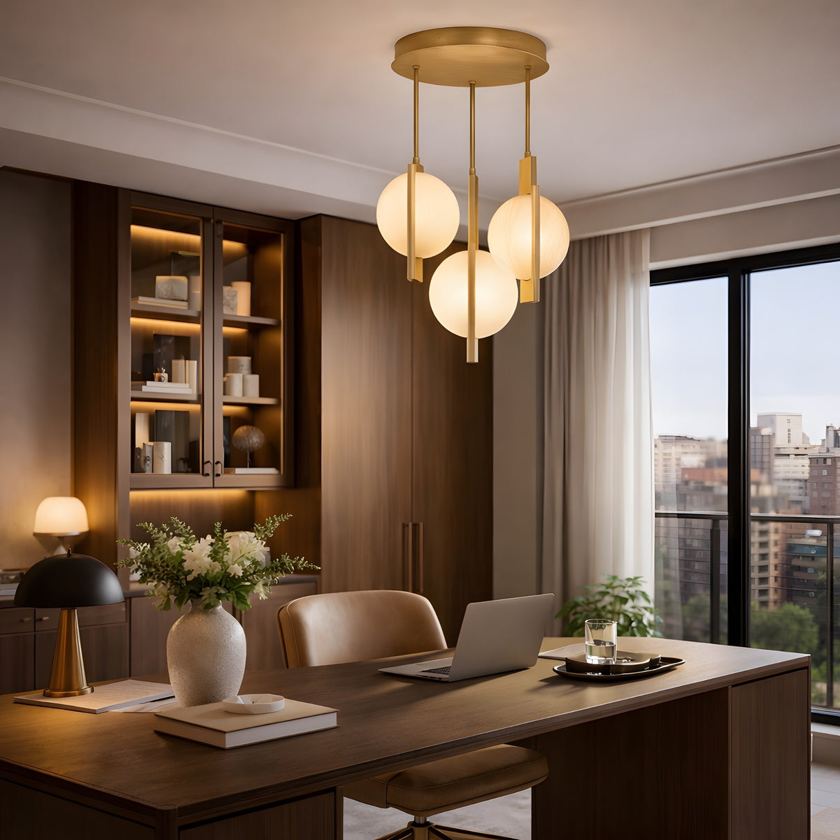 A modern home office with a large wooden desk, beige chair, and bookshelves features the Elyra 3 Light LED Alabaster Cluster Pendant in brushed brass. A city view window, flowers in a vase, and a laptop complete the scene.