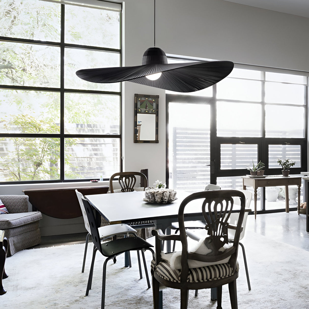 A modern dining area features the Eclipso 1 Light Pendant in black, a large dimmable fixture that hangs elegantly over a table set for four. Natural light pours through tall windows, highlighting the eclectic chairs and a potted plant centerpiece. The contemporary decor beautifully complements this bright and airy space.