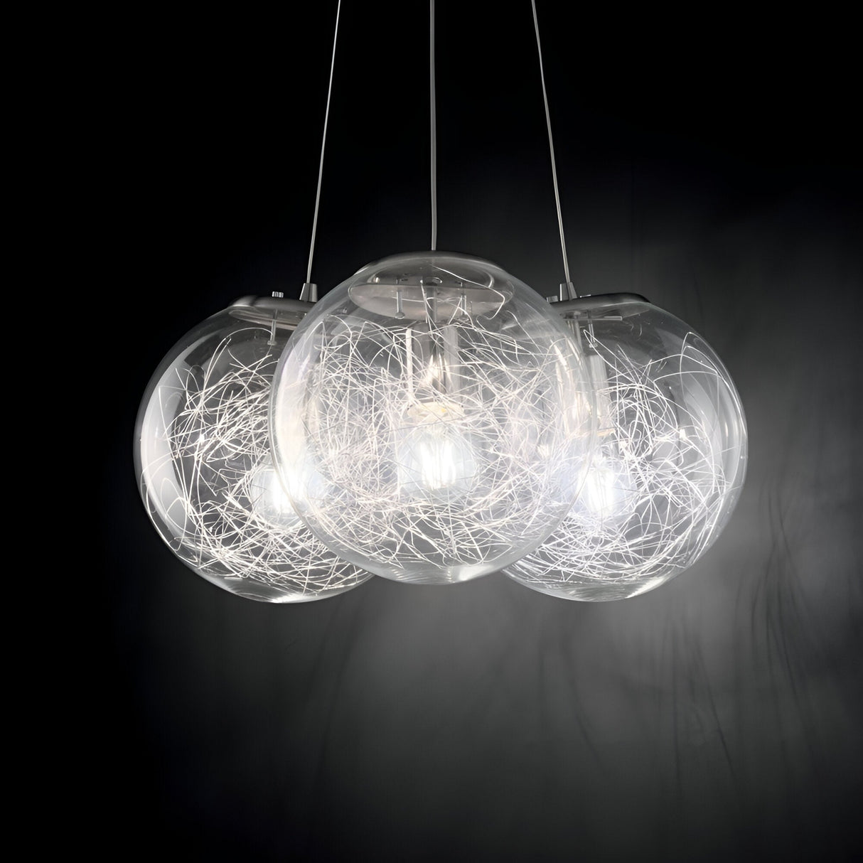 The Lyria 3 Light Cluster Pendant in silver showcases three clear blown glass diffusers, each enveloped by a decorative wire network surrounding a bulb. This elegant cluster pendant emits a soft, warm glow against a dark background, adding sophistication to any modern design lighting atmosphere.