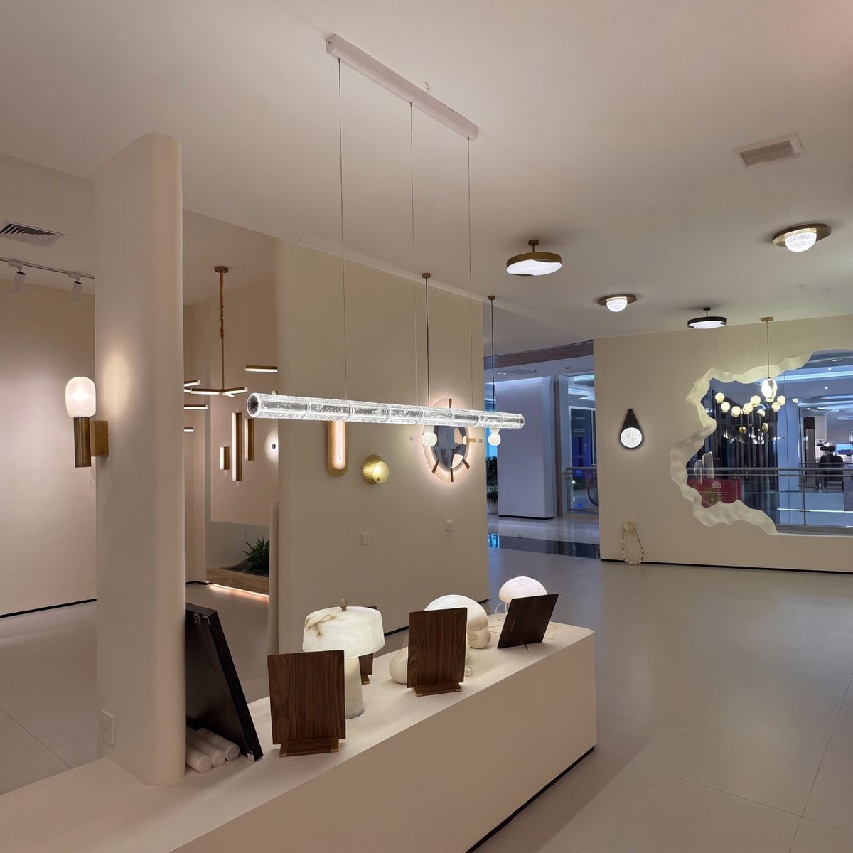 Liora LED Crystal Linear Chandelier 1.6m in chrome, segmented crystal tube suspended on cables, shown lit above a showroom display of lighting and decor pieces.