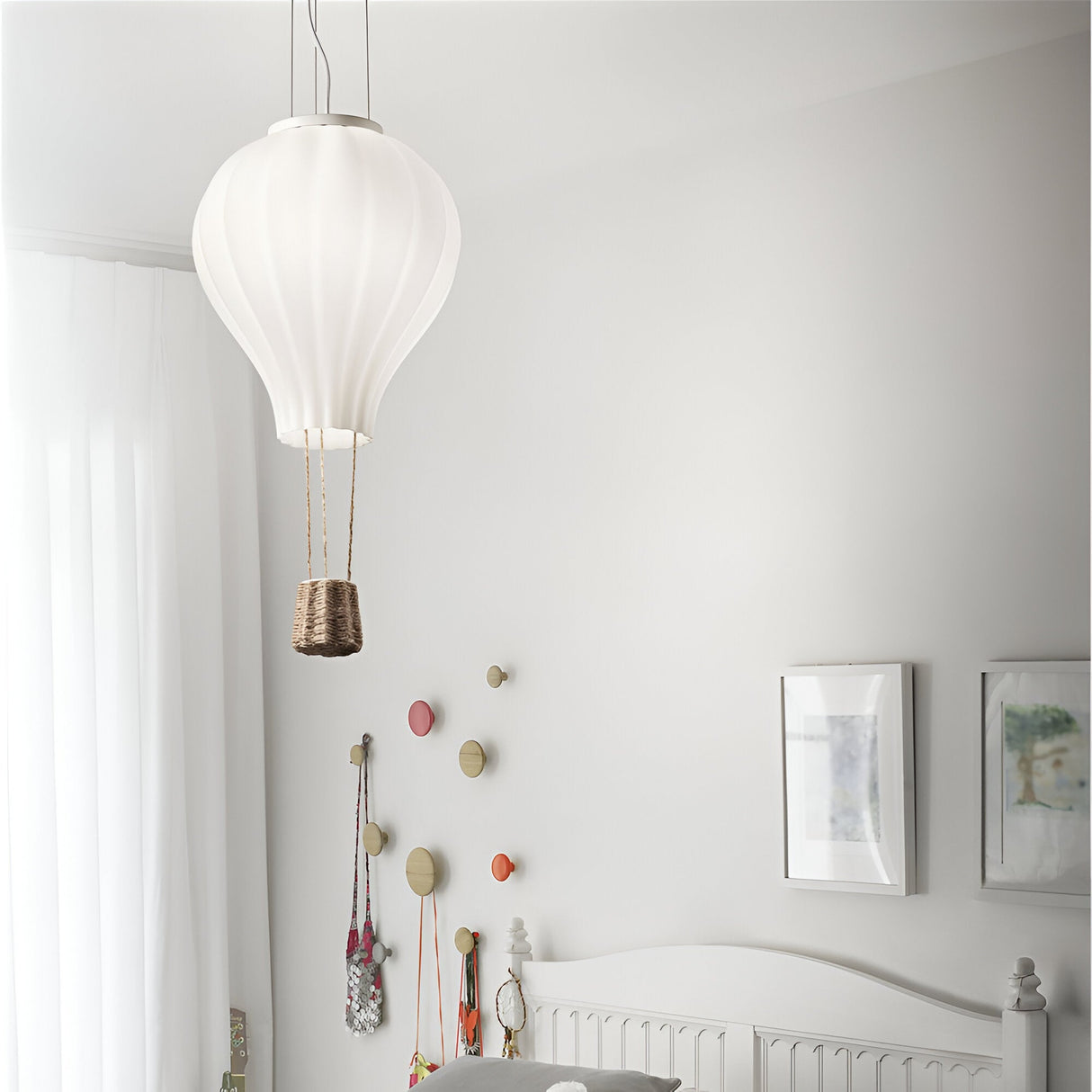 A minimalist bedroom features the Brilliance 1 Light Glass Pendant in a hot air balloon design, casting an ambient glow. The white walls, bed with a light-colored headboard, and colorful wall hooks enhance its charm. Framed art decorates the room as natural light filters through sheer curtains, highlighting its minimalist appeal.