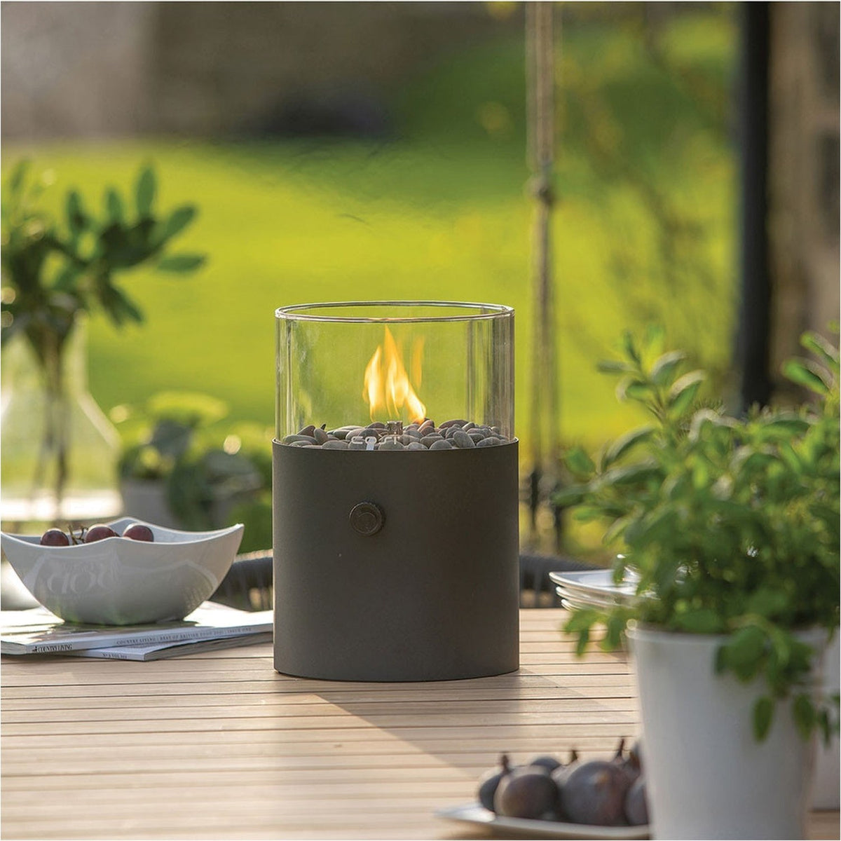 Cosi Extra Large Black Gas Flame Lantern, 30cm, with cylindrical glass top and black steel base holding pebbles and a lit flame, shown on an outdoor table setting.