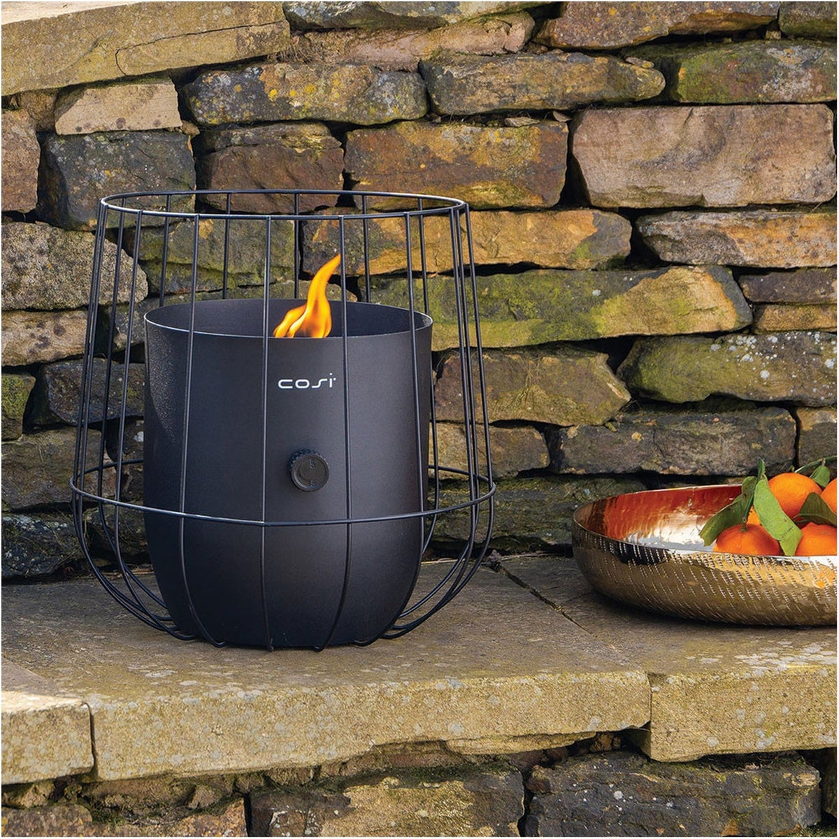 Cosi Basket Black Gas Flame Lantern, 31cm, with wire cage design and lit flame, shown outdoors on a stone ledge against a rustic stone wall.