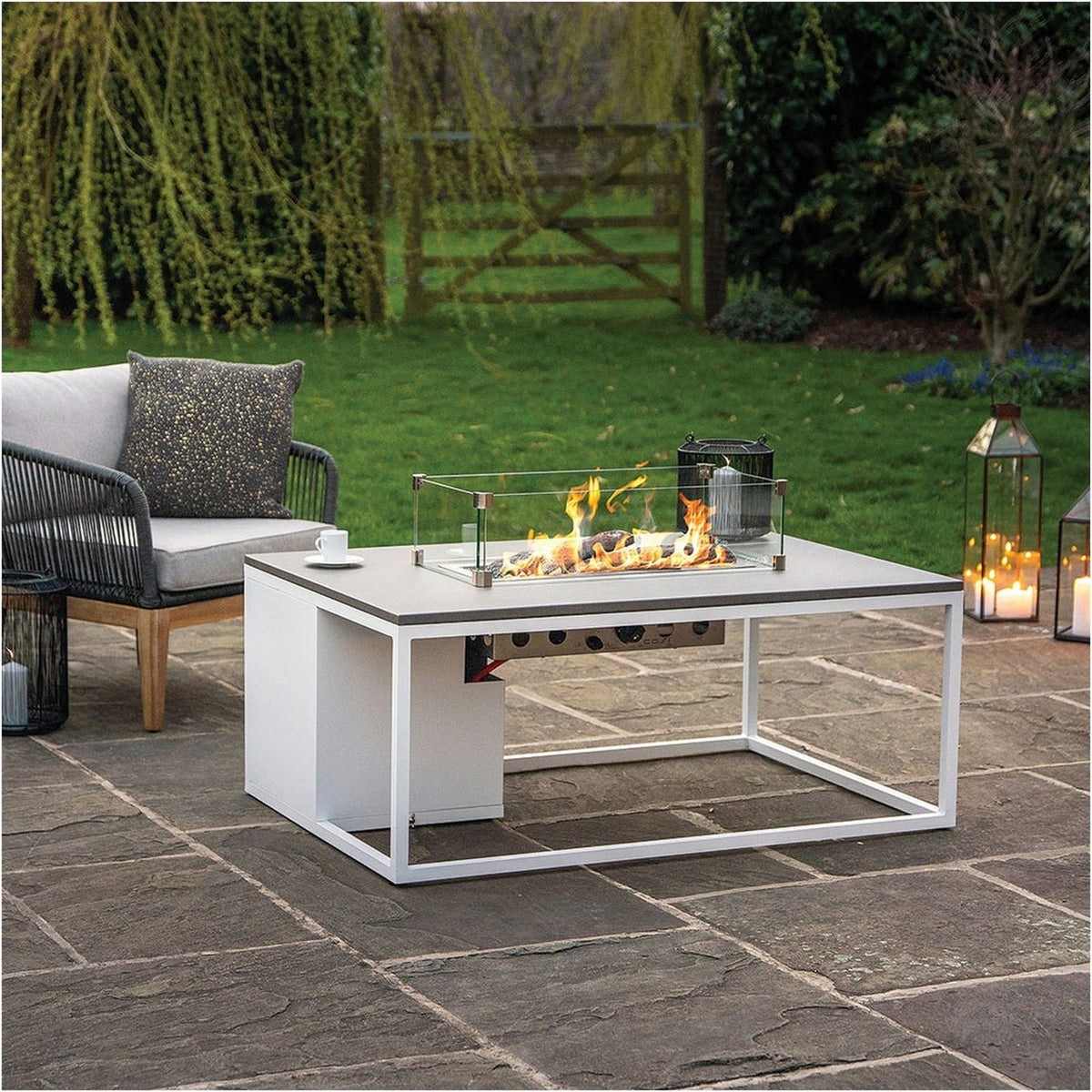 Cosi White & Grey Gas Fire Pit Table, 120cm, with glass windguard and lit burner, set on a patio beside outdoor seating and lanterns.