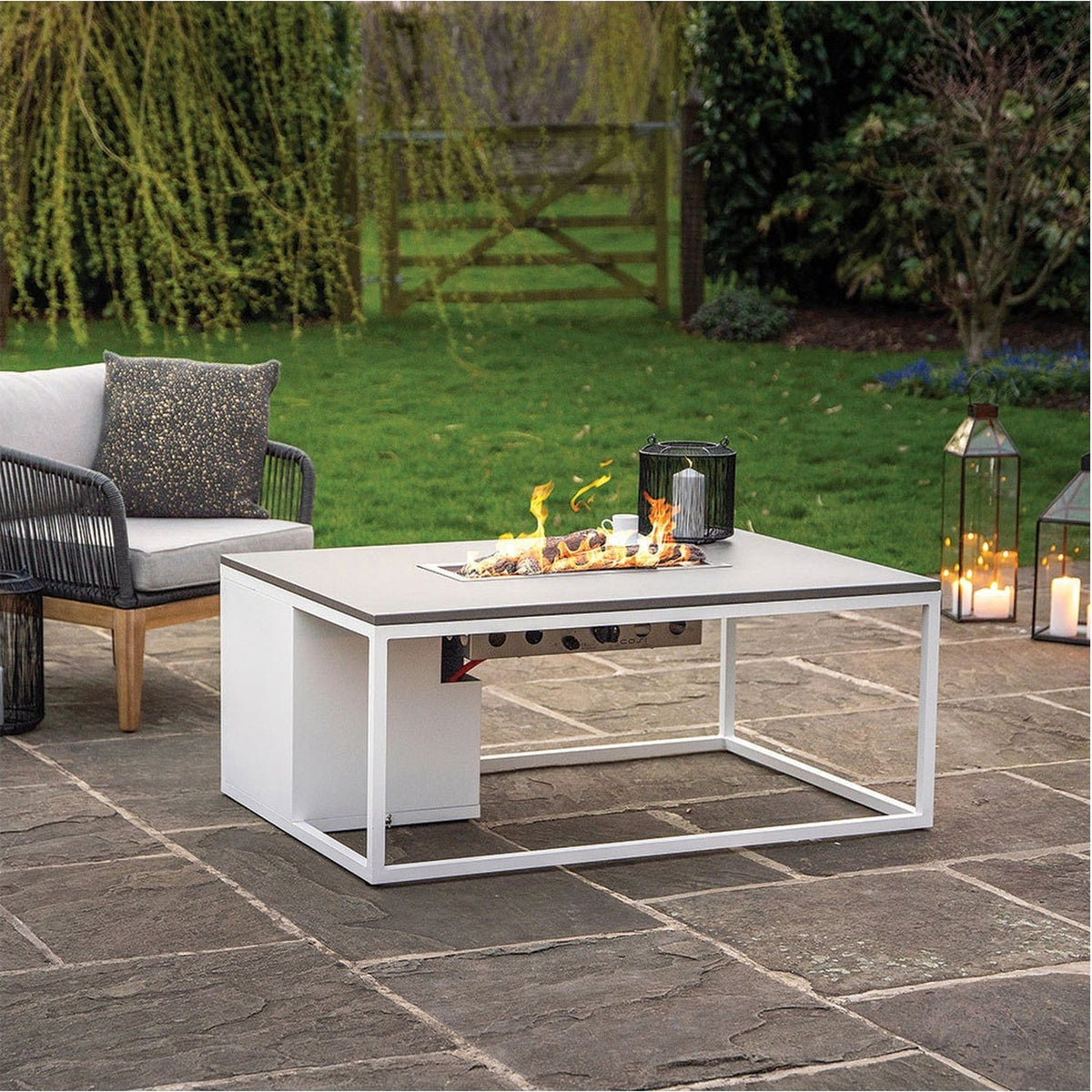 Cosi White & Grey Gas Fire Pit Table, 120cm, with lit burner and grey tabletop, white metal frame, shown on a garden patio beside outdoor seating and lanterns.