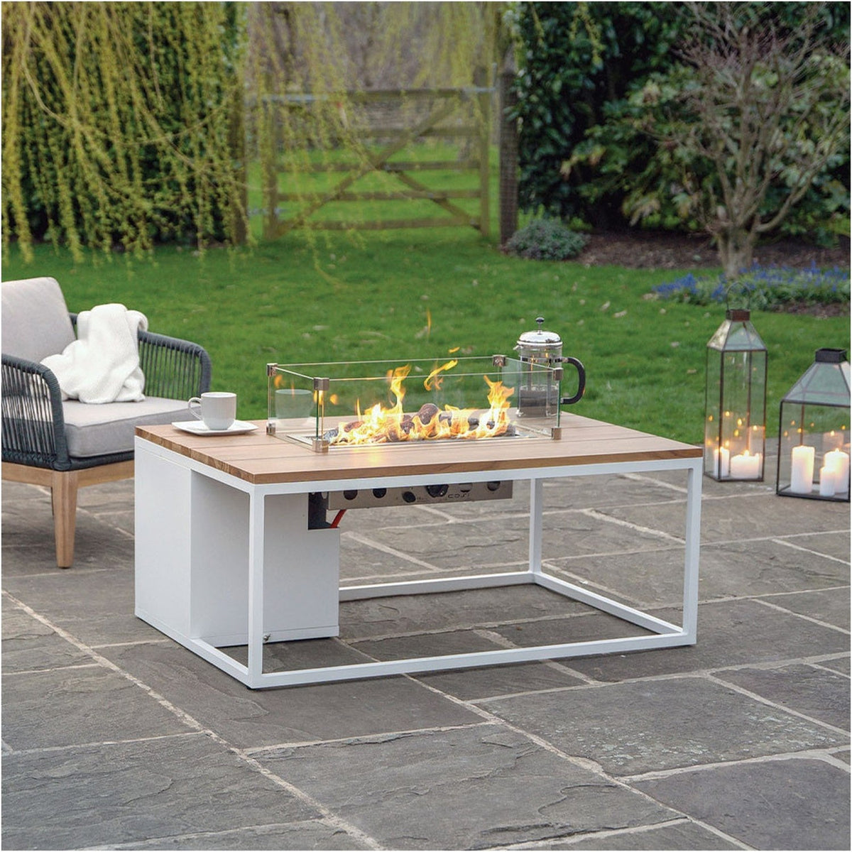 Cosi White & Teak Gas Fire Pit Table, 120cm, with white aluminium frame and teak-effect top, lit glass-guarded flame, shown on a patio beside a grey outdoor sofa and lanterns.