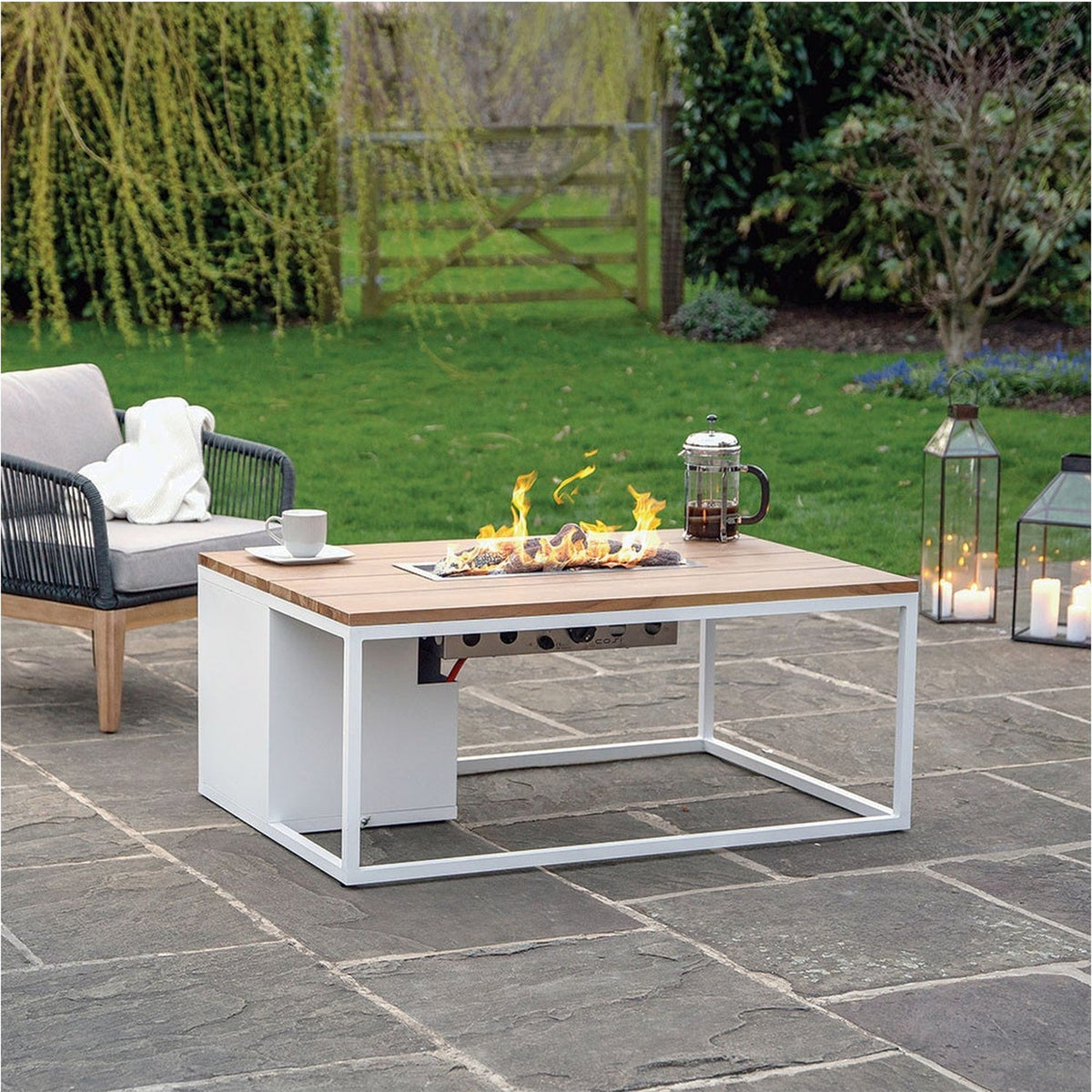 Cosi White & Teak Gas Fire Pit Table, 120cm, with white metal frame and teak-effect top, lit burner in a garden setting beside a sofa and lanterns.
