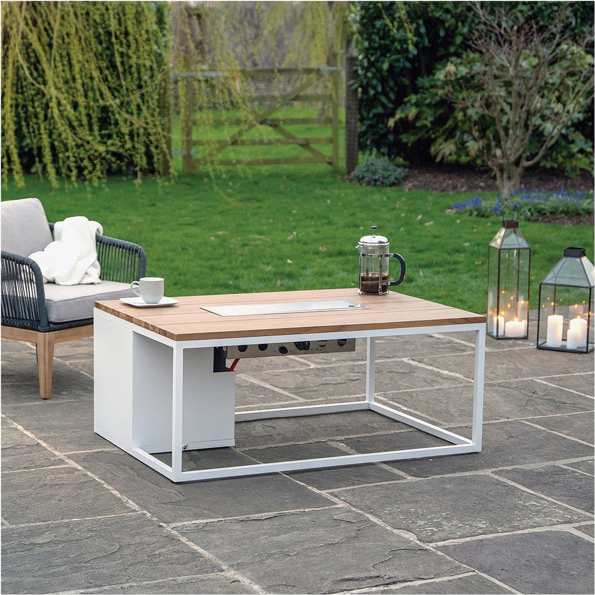 Cosi White & Teak Gas Fire Pit Table, 120cm, with white aluminium frame and wood top over a stainless steel burner, shown on a garden patio beside a sofa and lanterns.