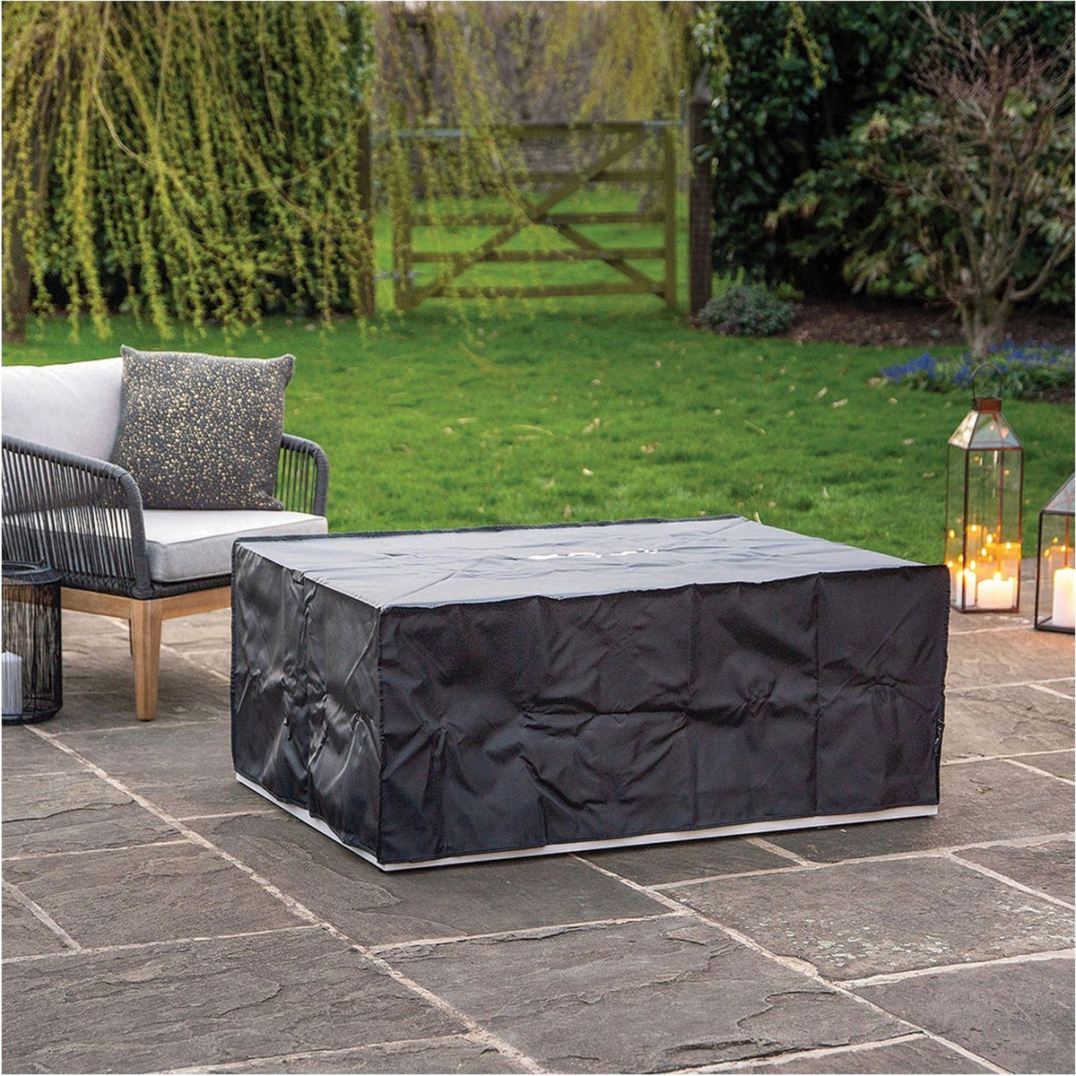 Cosi White & Teak Gas Fire Pit Table, 120cm, shown fitted with a black protective cover on a patio beside a grey outdoor sofa and lanterns.