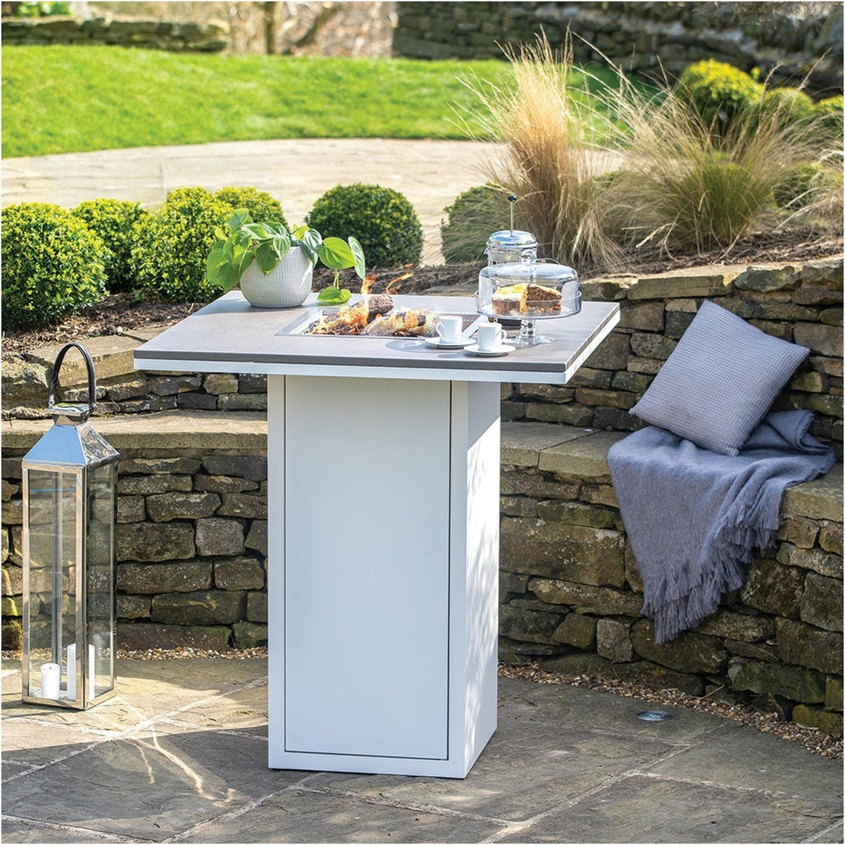 Cosi White & Grey Gas Fire Pit Bar Table, 100cm, square white base with grey tabletop and central fire pit, set on a garden patio beside a stone wall.