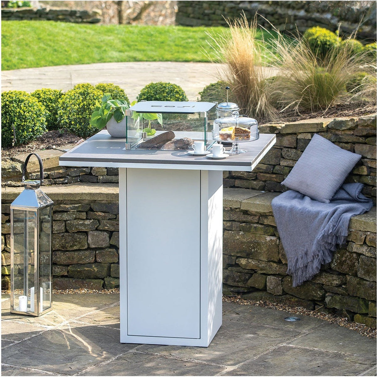 Cosi White & Grey Gas Fire Pit Bar Table, 100cm, with square grey top and white pedestal base, shown outdoors on a patio beside a stone wall with cushions.