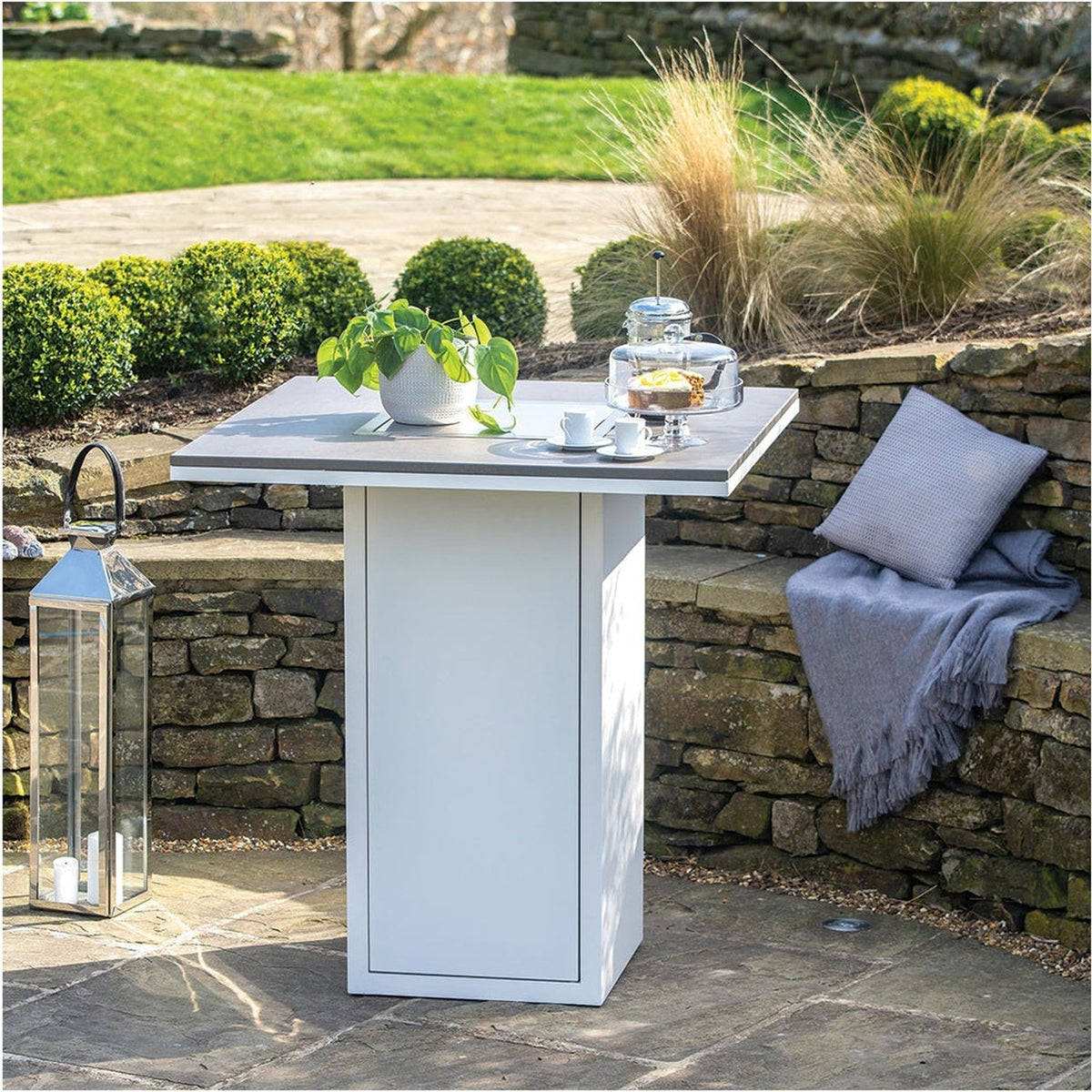 Cosi White & Grey Gas Fire Pit Bar Table, 100cm, with square grey top and white square pedestal base, shown on a garden patio beside a stone wall with cushions and a lantern.
