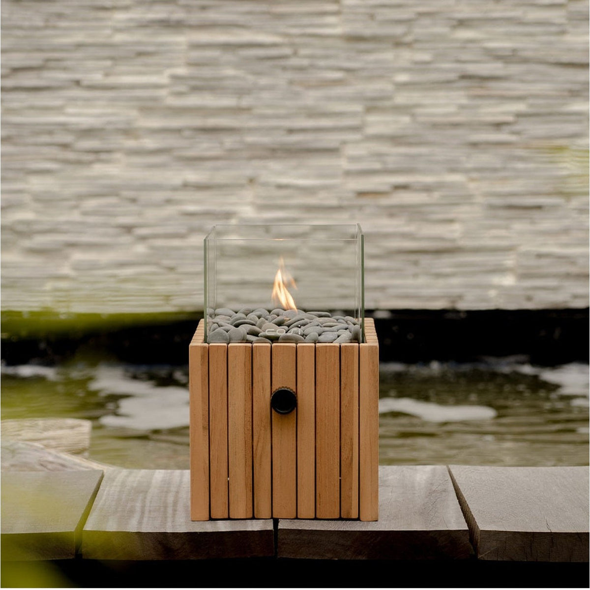 Cosi Timber Square Gas Flame Lantern, 30cm, with teak-look wood base and clear glass top over grey pebbles, lit flame, shown outdoors on a stone ledge by water.