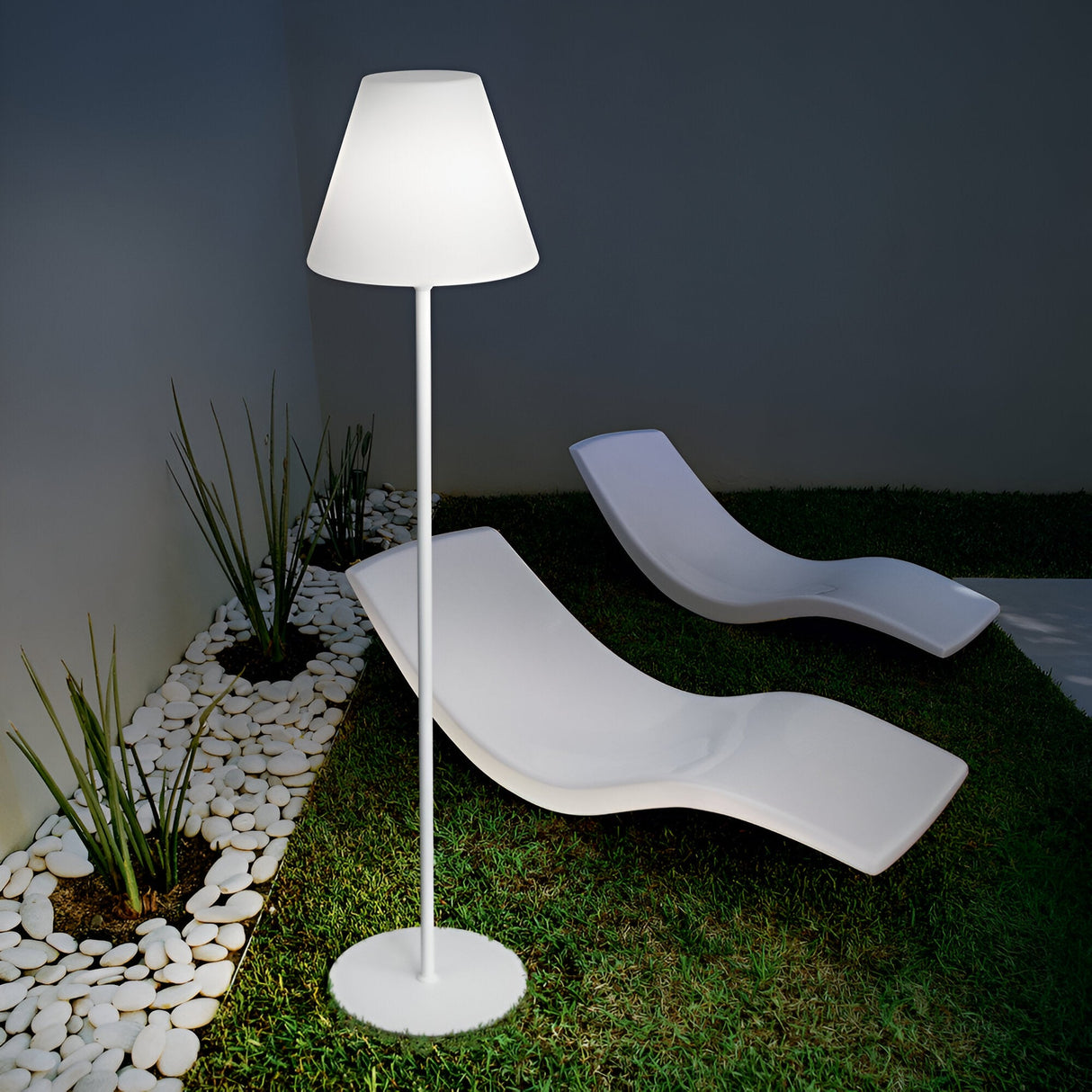 A minimalist outdoor setting at night showcases the Solora Outdoor Floor Lamp in white, casting light over two white curved lounge chairs placed on grass. The space is accented by a border of plants and white pebbles, creating an inviting and serene atmosphere.