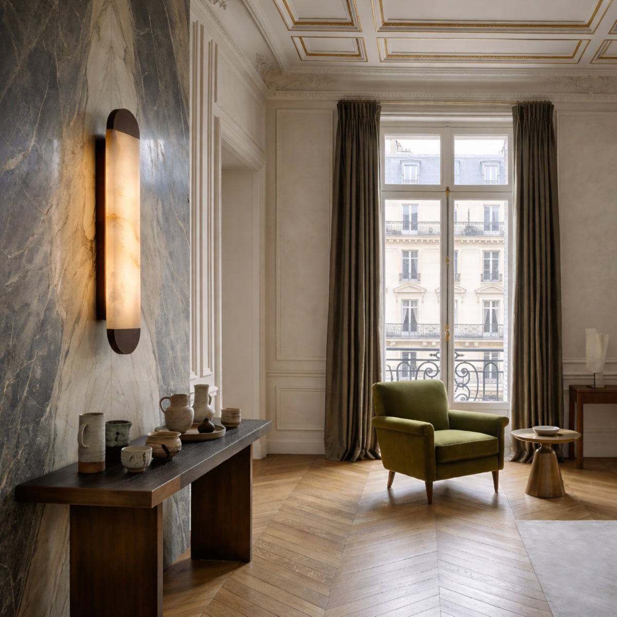 A stylish living room featuring a marble accent wall, the Orivelle LED Medium Vertical Alabaster Wall Light in brushed bronze, a wooden console with ceramics, green armchair, small round table, large window with heavy curtains, and Parisian views.