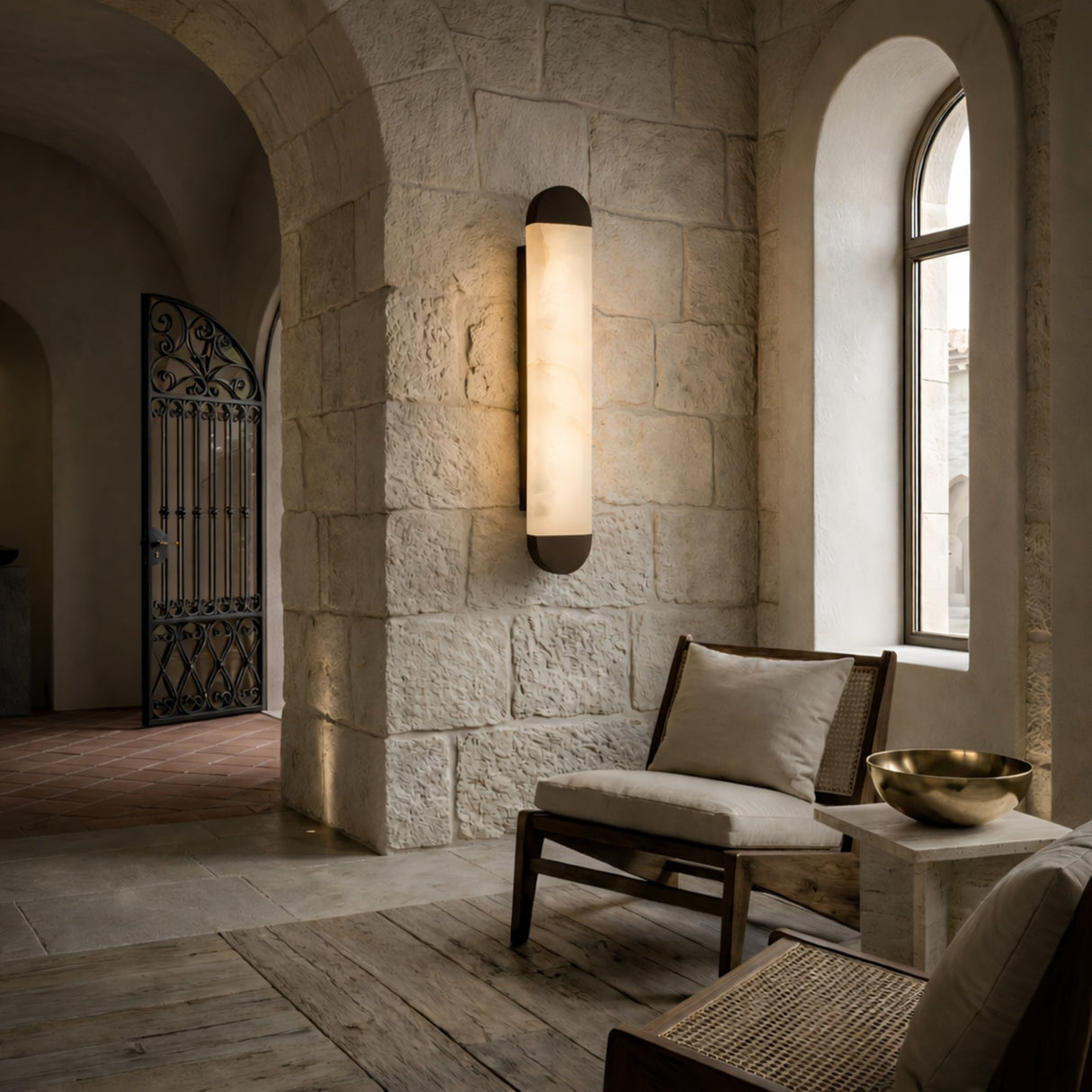 A cozy lounge area features a cushioned chair, side table, and the Orivelle LED Small Vertical Alabaster Wall Light in Soft White & Brushed Bronze, set against stone walls and arched windows for a peaceful, welcoming vibe.