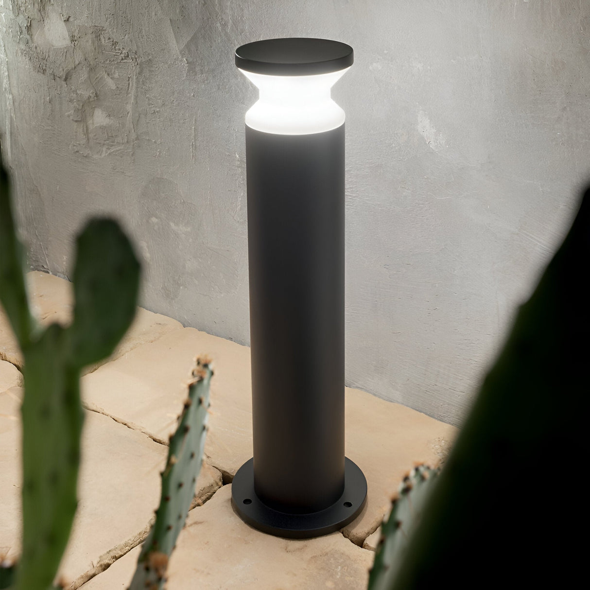 The Radiantix Outdoor Bollard Light - Black, 60cm boasts a modern aesthetic, featuring a cylindrical black base paired with a circular white illuminance that enhances the stone patio's ambiance. The foreground's cactus plants provide a natural contrast to its sleek design, and its IP44 rating ensures robust durability against various weather conditions.
