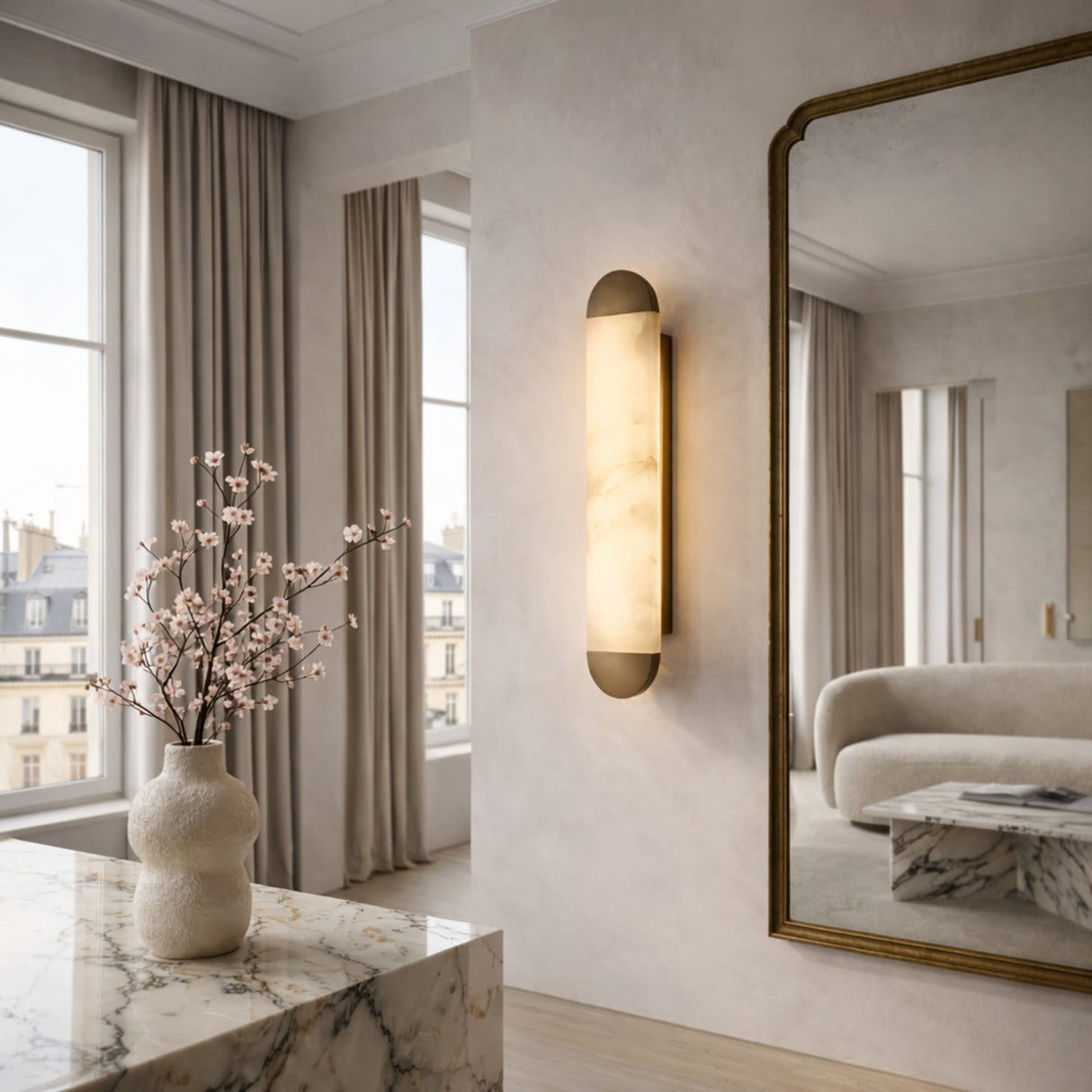 A modern, elegant living room with tall windows, beige curtains, a large wall mirror, a marble table with pink flowers, and the Orivelle LED Small Vertical Alabaster Wall Light in soft white and brushed bronze emits warm light.