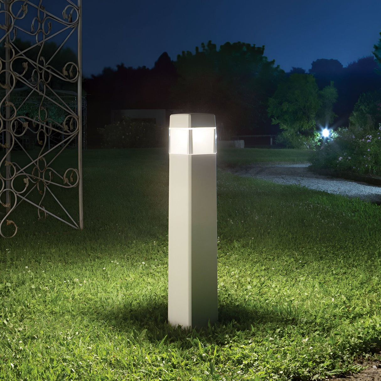 The Fluxen Outdoor Bollard Light - White, with its modern rectangular design, emits a bright light that illuminates the surrounding garden grass at night. In the background under the dark, clear sky, a winding path and an ornamental wrought iron structure can be seen. This aluminum lamp includes a dimmable GX53 bulb for customizable lighting.