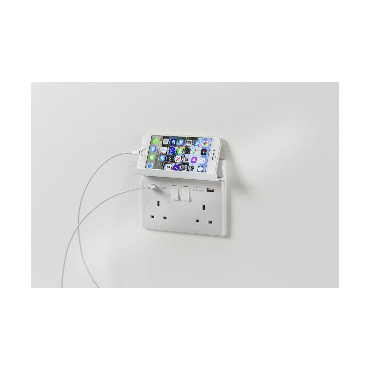 A smartphone is charging on a wall-mounted double switched socket with USB ports, resting securely on a 2 Gang Fold Away Phone Holder - White above the outlets. It is supported by a charging cable, all elegantly installed on a plain white wall.
