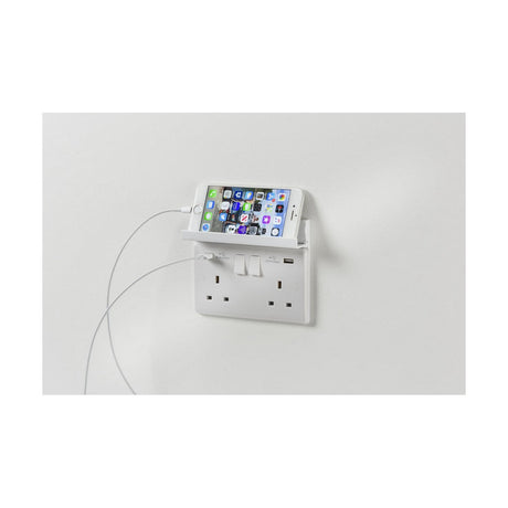 A smartphone is charging on a wall-mounted double switched socket with USB ports, resting securely on a 2 Gang Fold Away Phone Holder - White above the outlets. It is supported by a charging cable, all elegantly installed on a plain white wall.