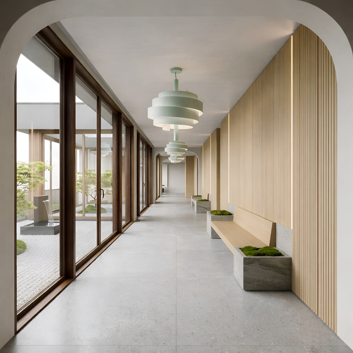 A modern hallway features large windows, light wood panel walls, gray benches with small planters, and a striking Picasso Pendant Light in green; through the windows, a lush courtyard with plants is visible.