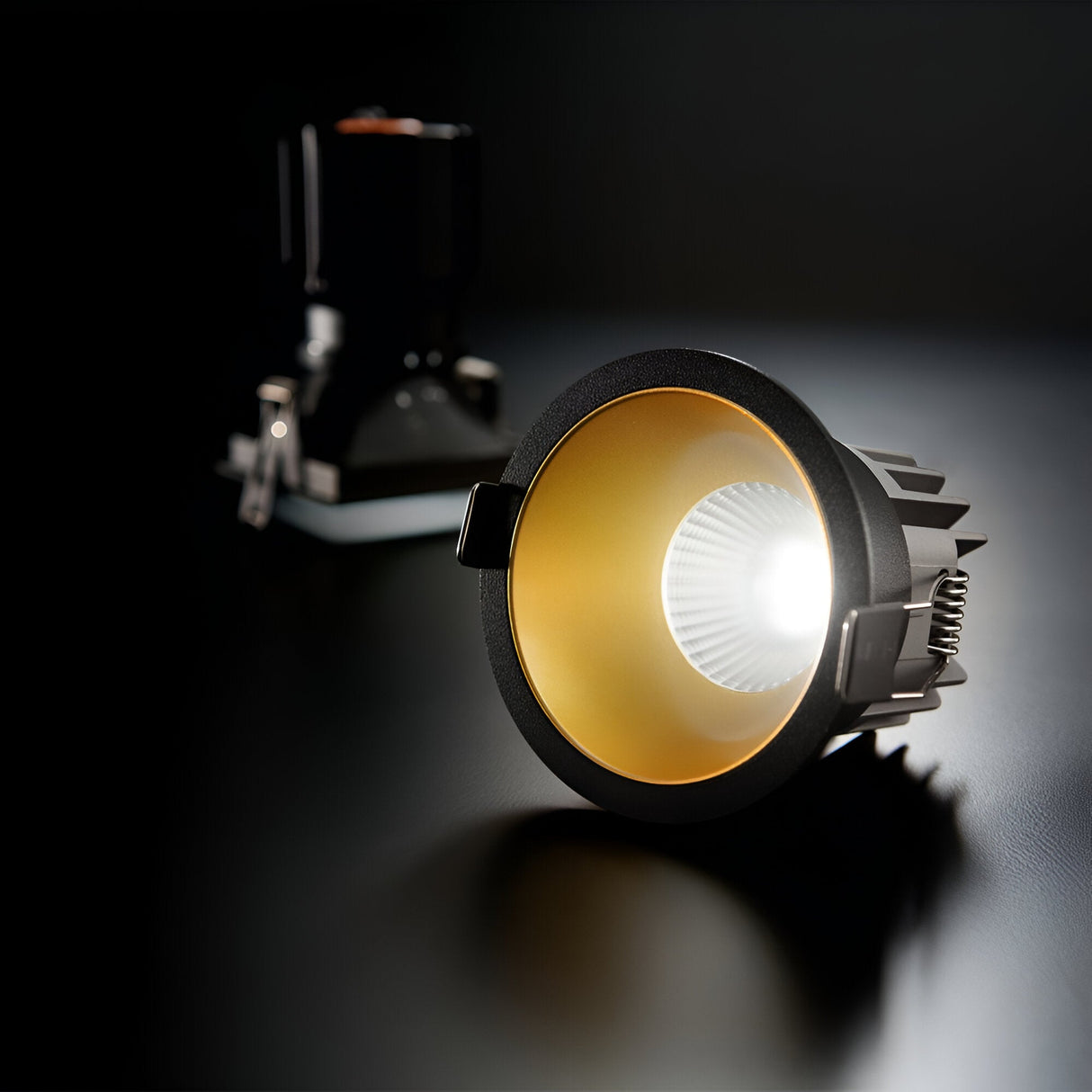 A close-up captures the Sally LED Round Recessed Trim Downlight, emitting a warm white light at 3000K. The fixture's sleek design features a gold and black finish, with another similar fixture blurred in the background. It showcases its advanced Chip on Board (COB) technology against a dark surface.