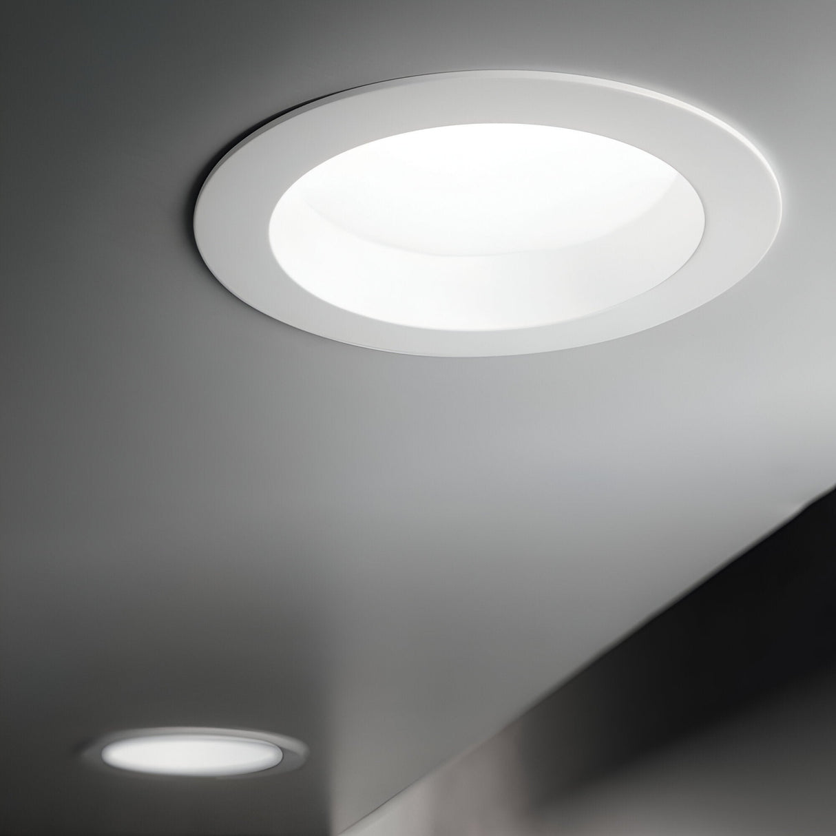 A modern ceiling with two Radiara-2 Round LED Recessed Downlight 71mm, each offering IP44 protection, casts a gentle 3000K white glow against the smooth gray surface, resulting in a minimalist and sleek aesthetic.
