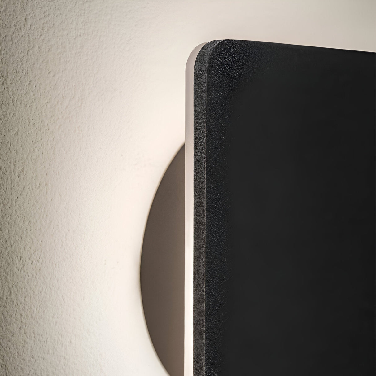 Close-up of a NovaLite LED Square Ceiling & Wall Light, featuring a circular base and a rectangular black panel. Soft, energy-efficient light shines from behind the panel, casting a subtle glow on the wall.