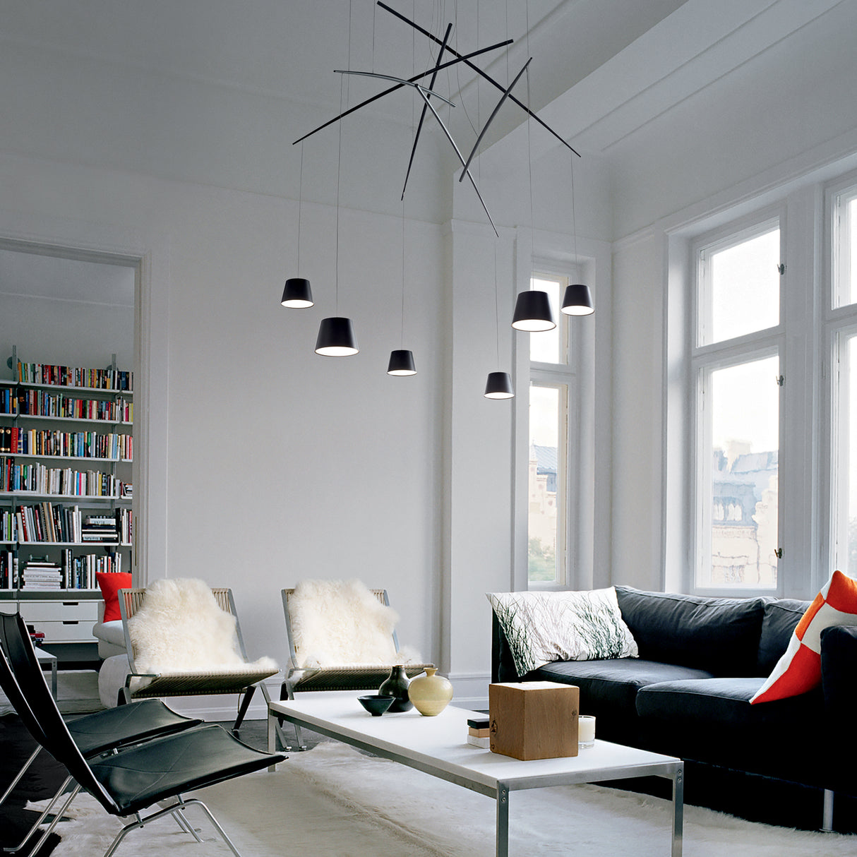 A modern living room with a black sectional sofa, white coffee table, and the sleek Illumia 6 LED Light Suspension 3000K 37W in black. The room features large windows letting in natural light, a bookshelf filled with books, and chairs with fur cushions.