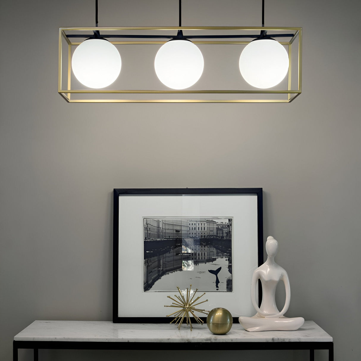 A modern interior showcases the Dawn 3 Suspension Light in brass and black with three spherical bulbs above a table. The table exhibits a framed black and white photo, a sleek abstract sculpture, and a starburst ornament, all accentuated by the chic contrast of brass and black tones.