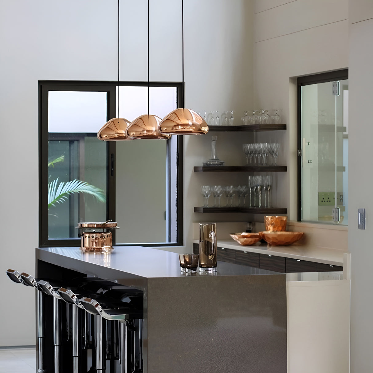 A modern kitchen showcases a sleek gray island accompanied by black bar stools and the energy-efficient Gulf Glass Pendant Light in copper. On the countertop, there is a coordinating copper pitcher and bowl, while open shelves exhibit glassware against a white wall. A large window allows for an expansive view of the greenery outside.