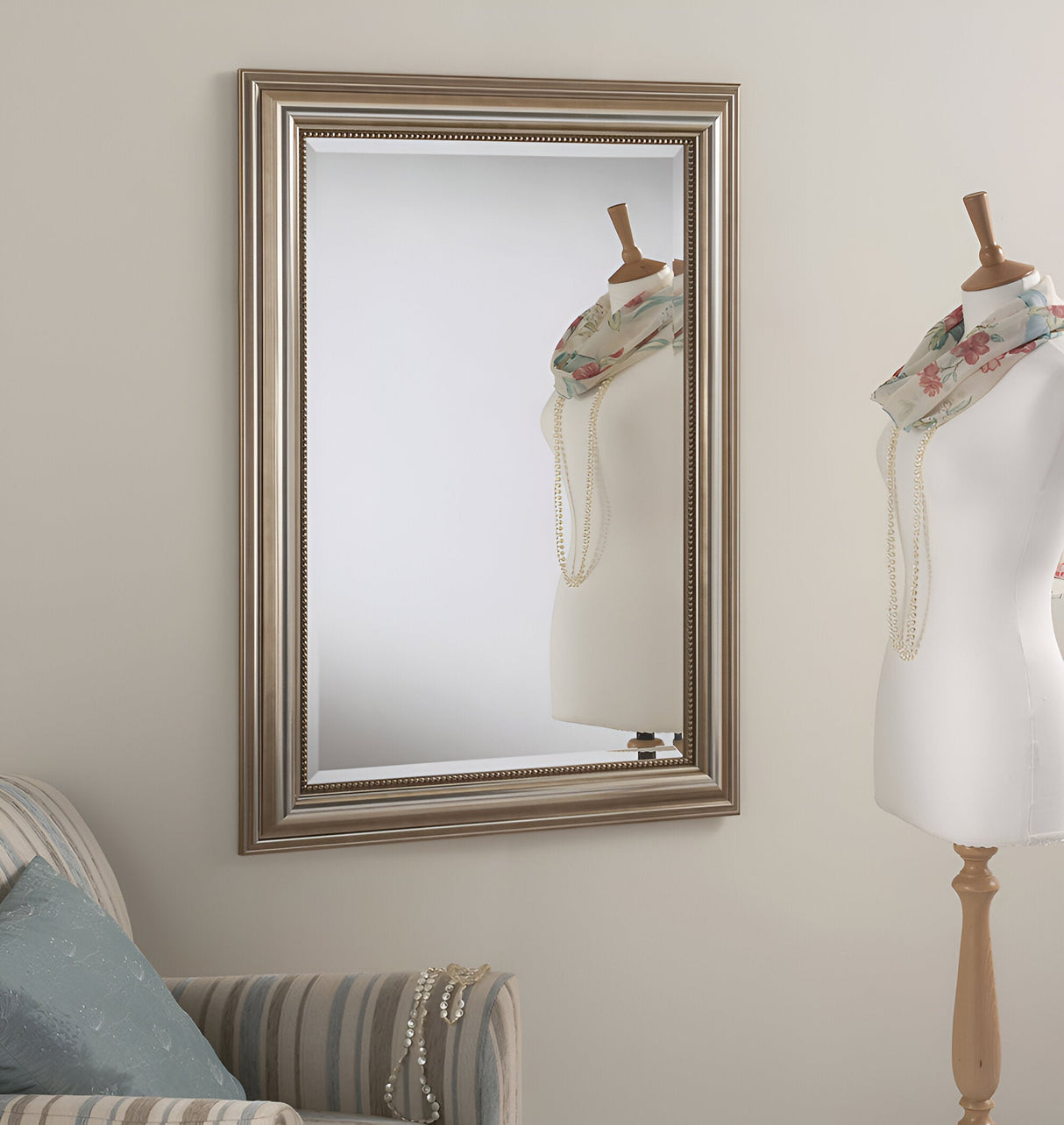 An elegant room decor showcases a mannequin with a floral scarf and pearl necklace next to the Verity mirror, which features a large rectangular silver frame. A striped armchair with a blue pillow and draped pearls peeks in from the left.