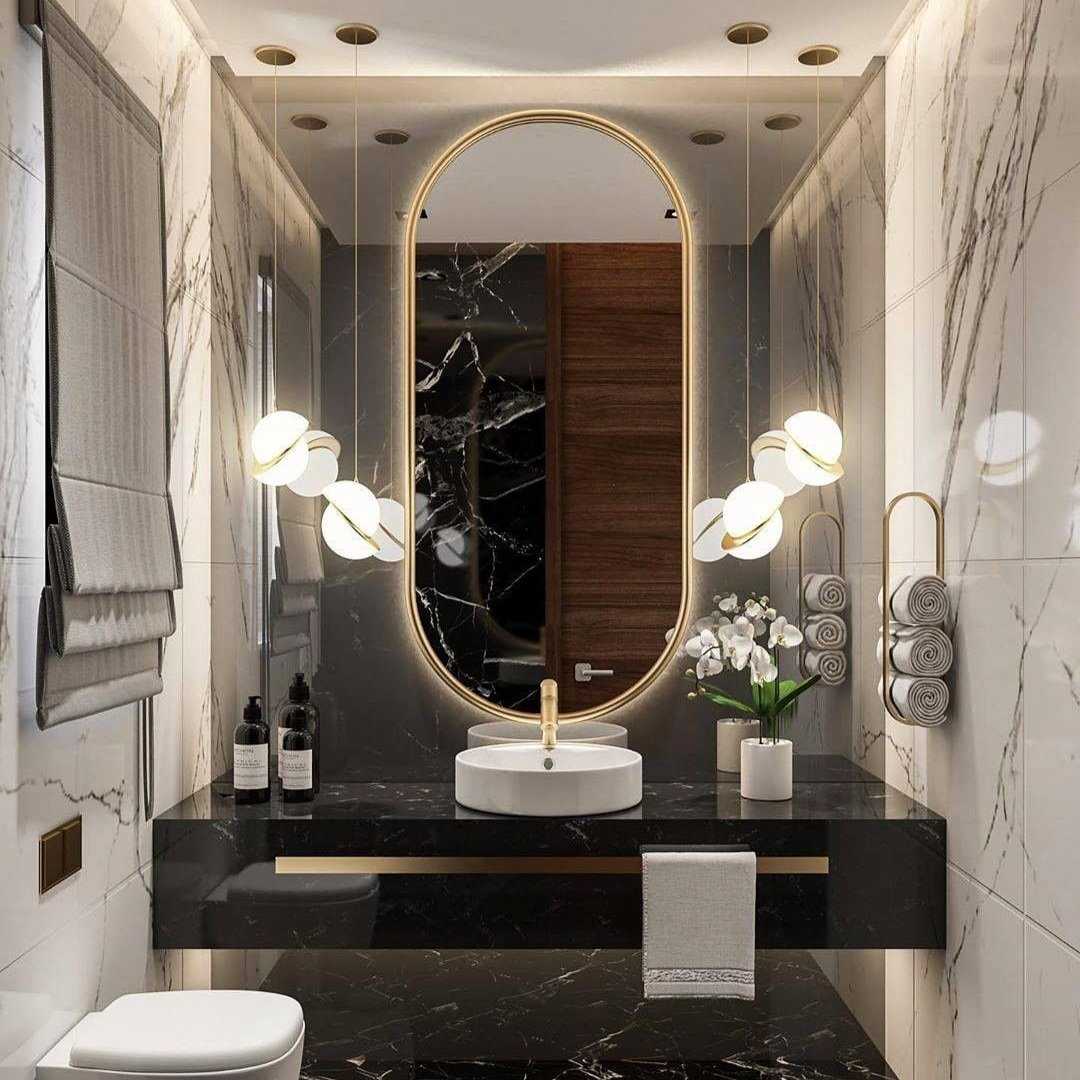 This modern bathroom showcases a contemporary design, highlighted by a large oval mirror surrounded by the golden glow of the Arc Pendant Light in Gold & White. It features a black marble countertop with a white sink, brushed brass accents, decorative lighting, rolled towels, and a white flower arrangement set against marble walls.
