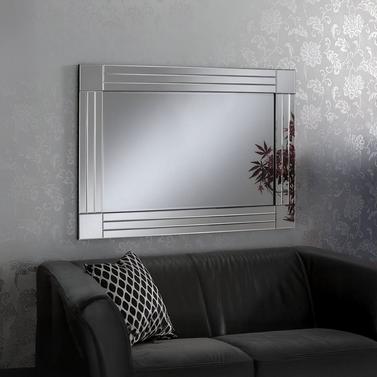 The Aurelia Rectangle Wall Mirror Clear (120x80cm) hangs on a patterned gray wall above a dark sofa. A black-and-white cushion adds elegance, while the mirror’s reflection captures the greenery of nearby plants.
