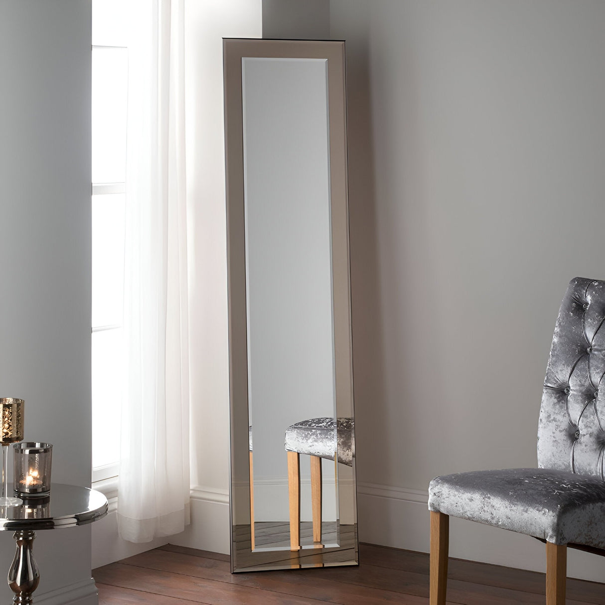 A modern accent, the Silhouette Leaner Wall Mirror Bronze (181x44cm) stands against a light gray wall beside sheer white curtains, paired with a gray tufted chair and a small round table topped with a candle.