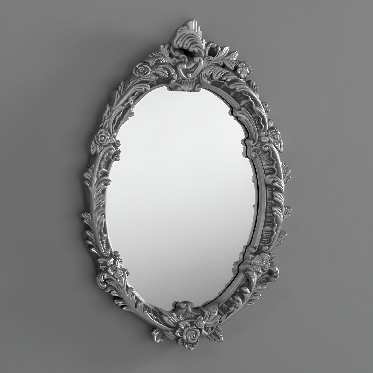 The Mirage Oval Wall Mirror Silver (64x46cm) features a floral-patterned frame, bringing decorative depth and charm to any plain gray wall.