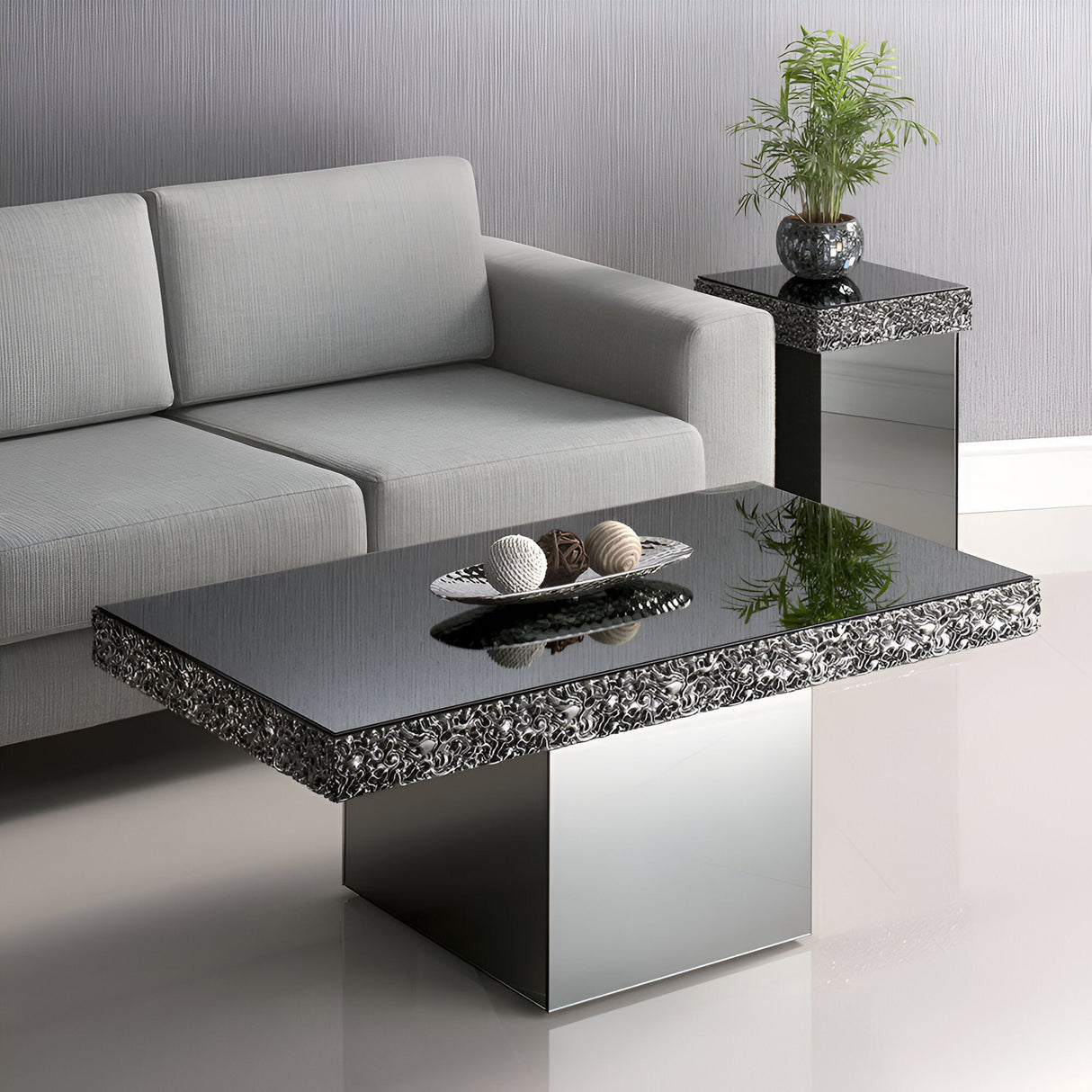 A modern living room showcases a gray sofa, the Stratos Mirrored Coffee Table - Grey with decorative edges and a bowl of decorative balls, plus a matching side table featuring a plant.