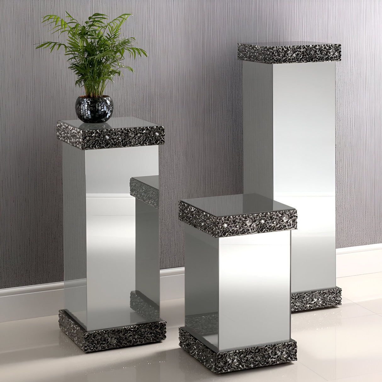 Three Pavilion Mirrored Plinths in smoked grey with black trim stand on a shiny tiled floor. The tallest pedestal holds a leafy green plant in a black vase, set before a modern textured gray wall.