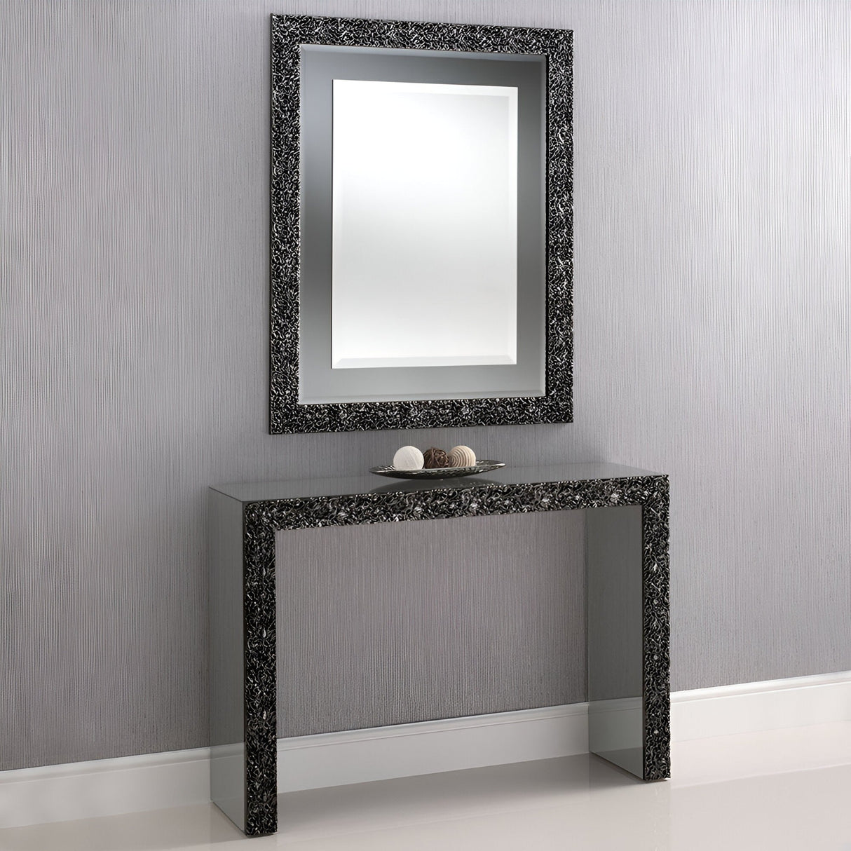 The Pavilion Mirrored Console Table - Smoked Grey, featuring reflective panels, stands against a light gray textured wall. It’s paired with a matching framed mirror and tray of décor, highlighting the rooms modern minimalist style.