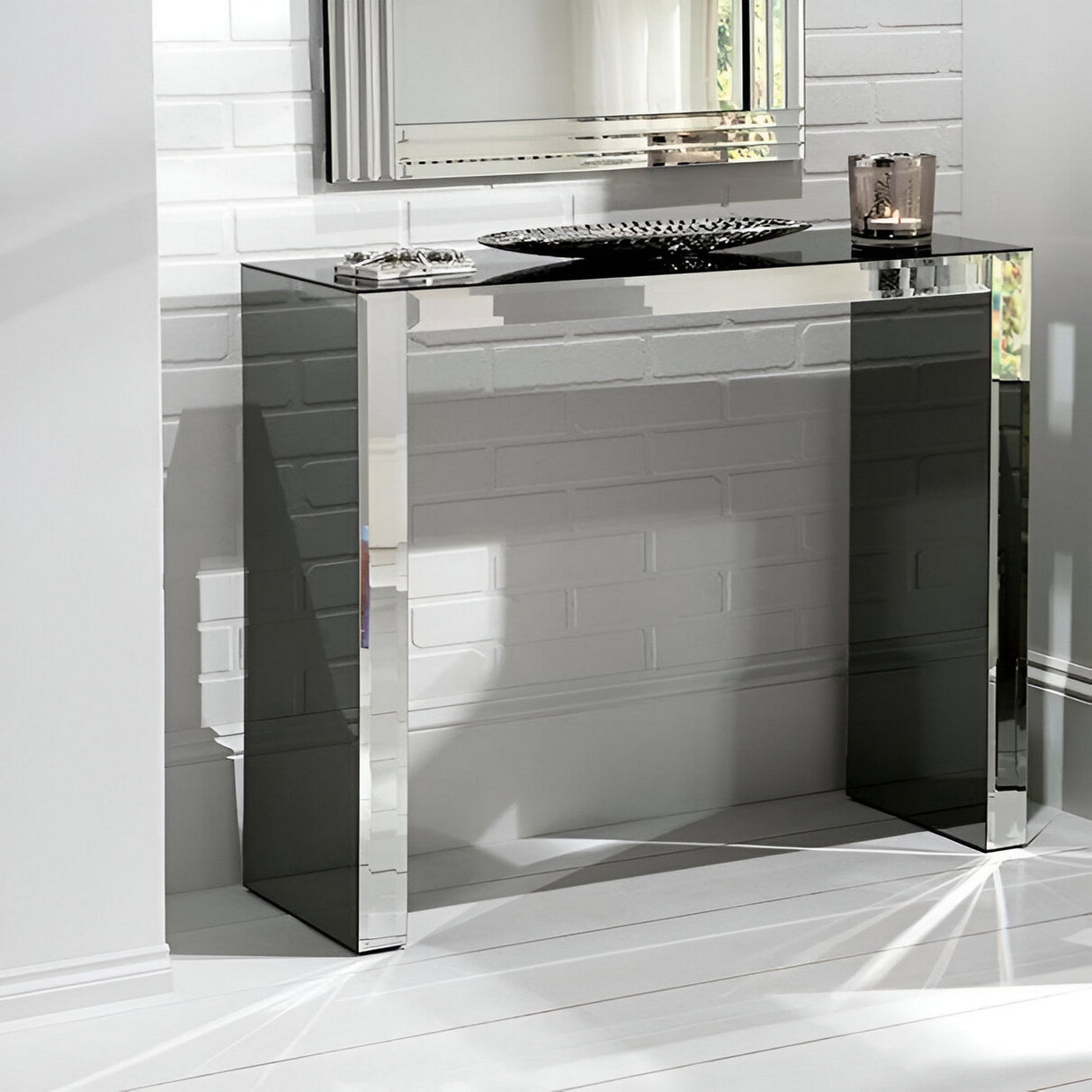 The Atrium Mirrored Console Table - Grey stands against a white brick wall, its reflective surface topped with a silver tray, ornament, and candle holder. This table adds brightness and elegant style to any minimalist space.