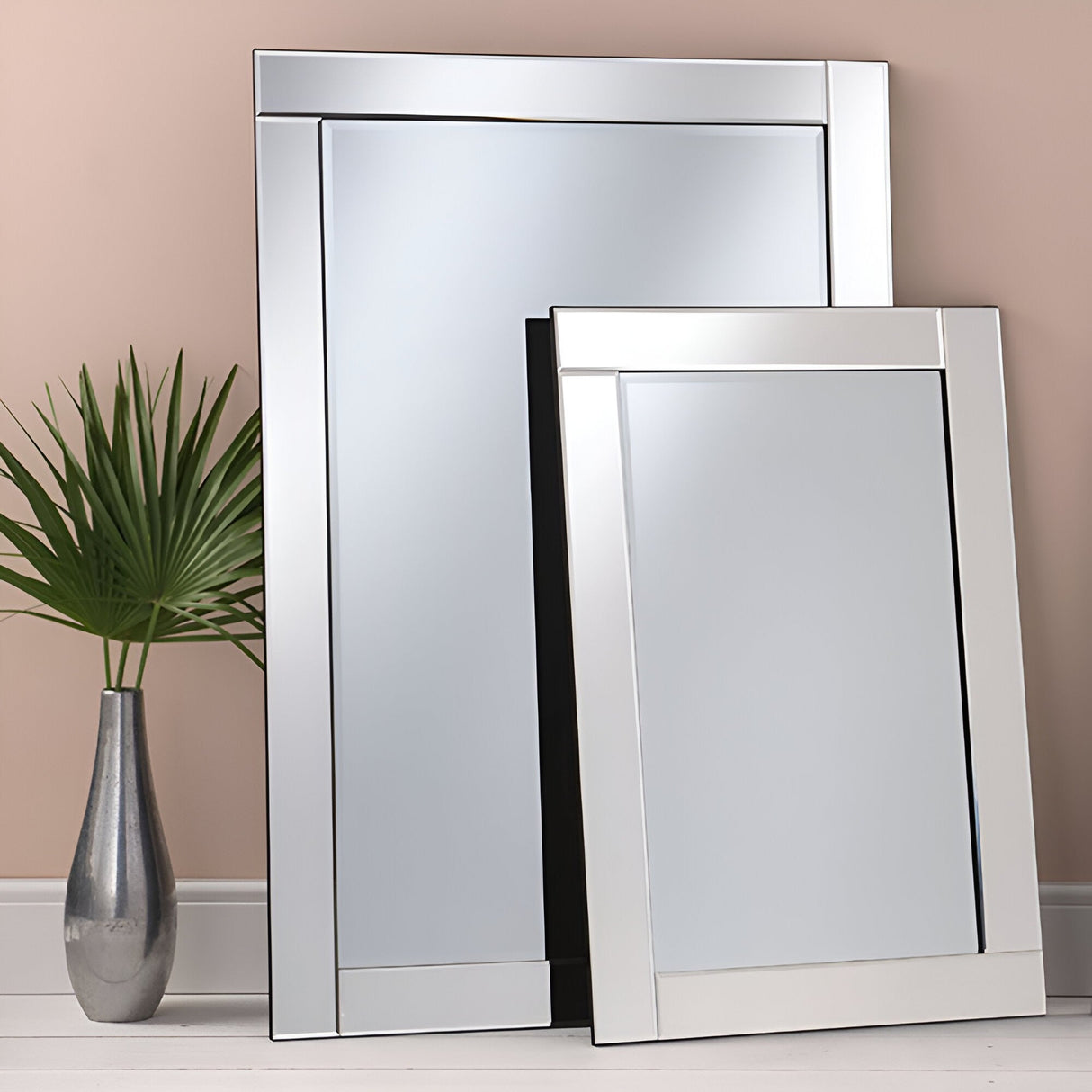 Two Aurelia Rectangle Wall Mirror Clears (90x60cm) with metallic frames lean against a beige wall on a white floor. A silver vase with a green palm leaf adds versatility, making this reflective setup an elegant statement piece.