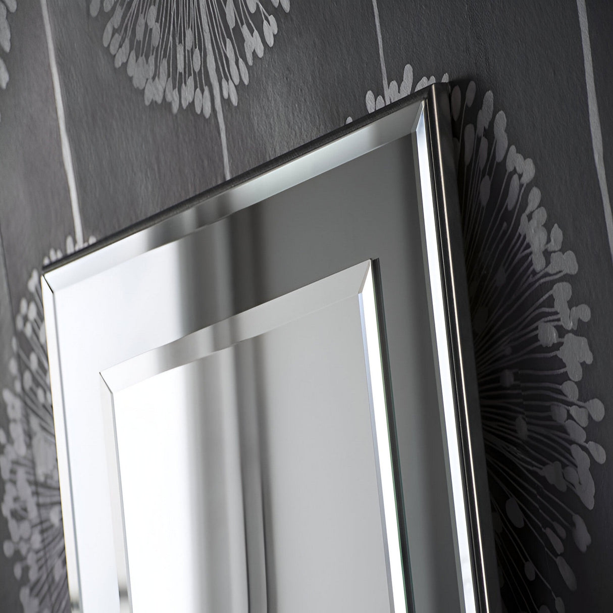 Close-up of the Focus Leaner Mirror - Grey, featuring a beveled, angled frame and contemporary design. The mirror reflects light and stands out against dark wallpaper with a large, abstract gray floral pattern.