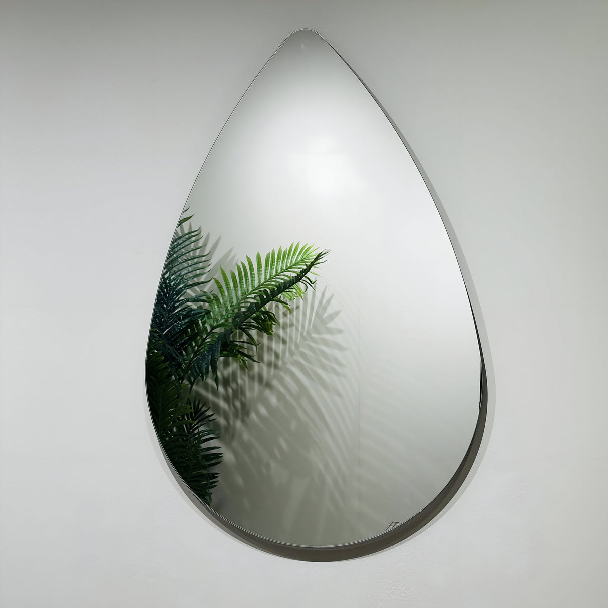 The Oculus Droplet Wall Mirror Clear (90x60cm), with its unique teardrop shape, is mounted on a white wall and reflects green fern leaves along with their shadows.