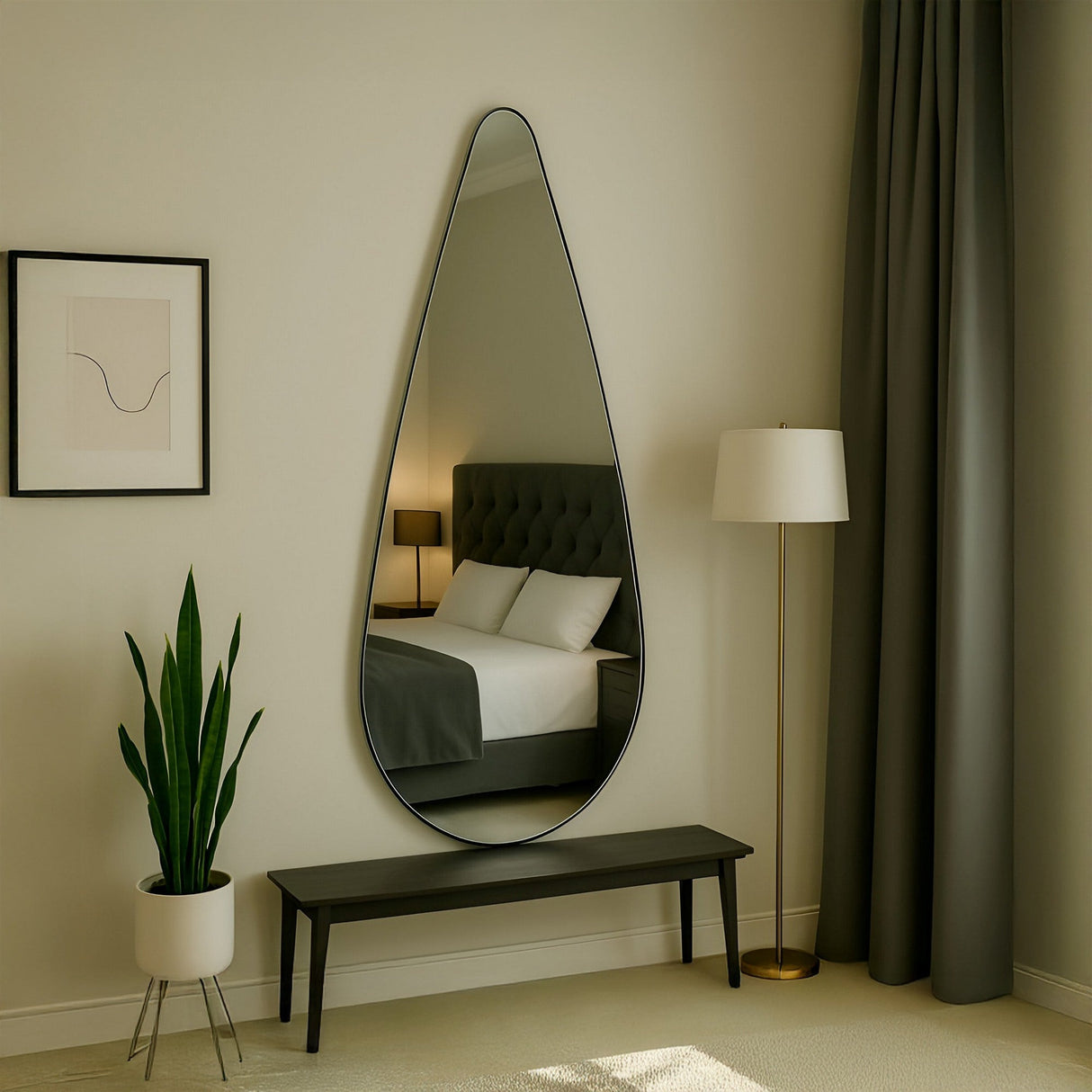 A modern bedroom showcases the Oculus Droplet Wall Mirror - Clear with a frameless design above a black bench, near a potted plant, gold floor lamp, abstract wall art, and a bed with a tufted grey headboard reflected in the mirror.