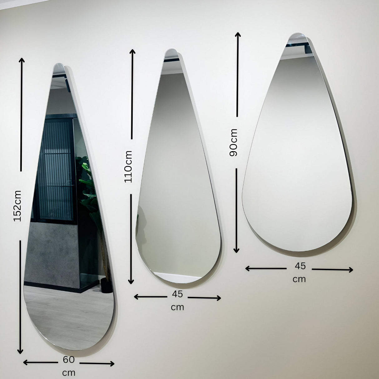 Three Oculus Droplet Wall Mirrors - Clear, each in a different size, are mounted on a white wall. Their frameless, teardrop design suits minimalist styles, and the largest mirror reflects part of a modern interior.