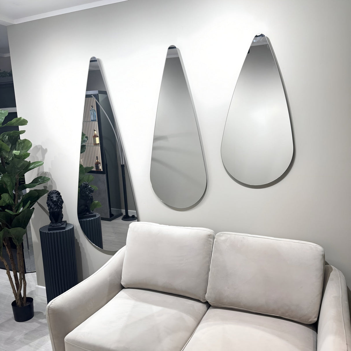 A modern living room with a light beige sofa, three frameless Oculus Droplet Wall Mirror - Clear pieces, a tall plant, and a black pedestal topped with a small sculpture.