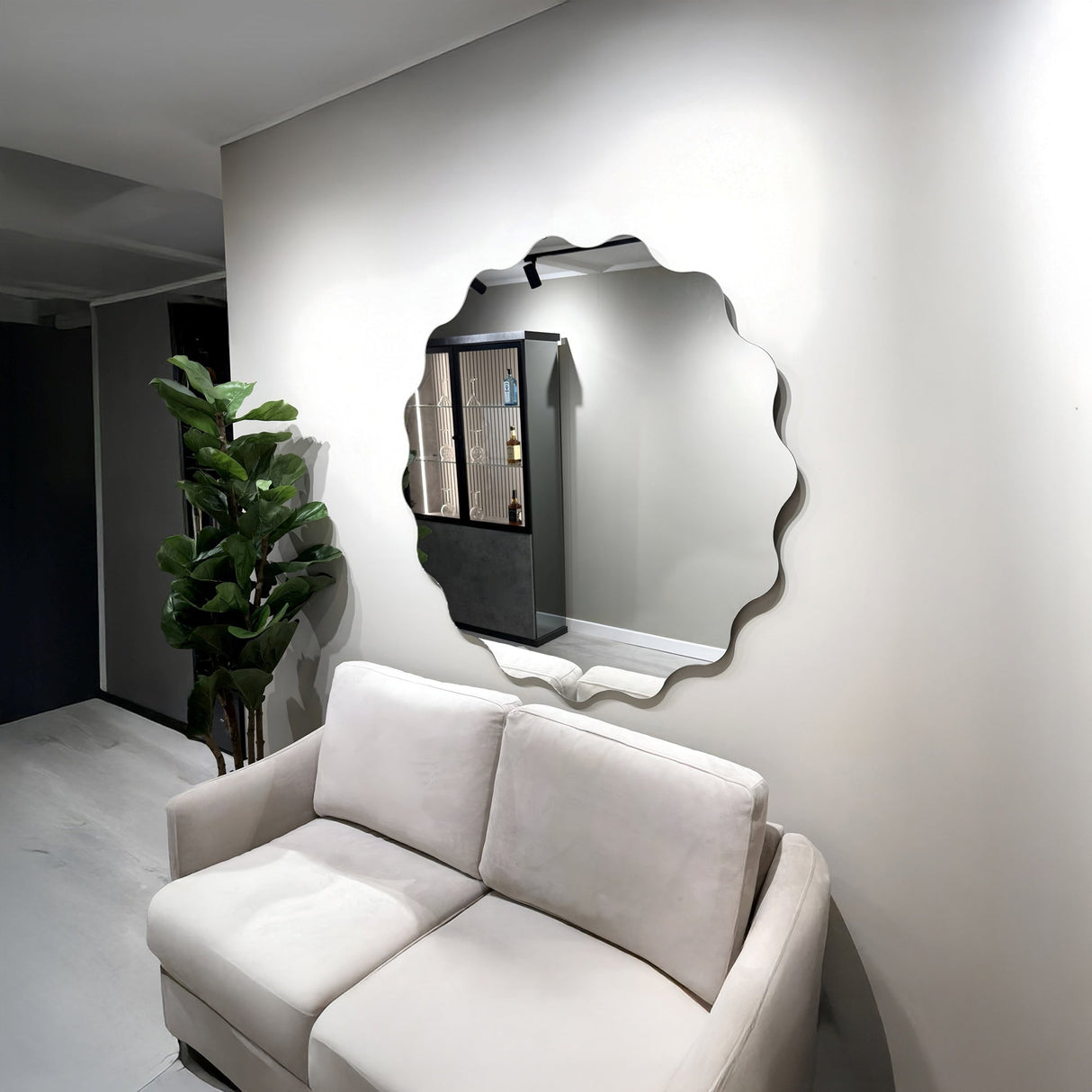 A modern living room with a beige sofa, a tall green potted plant, and the Lumeo Circle Wall Mirror - Clear on a light gray wall reflecting a shelving unit.