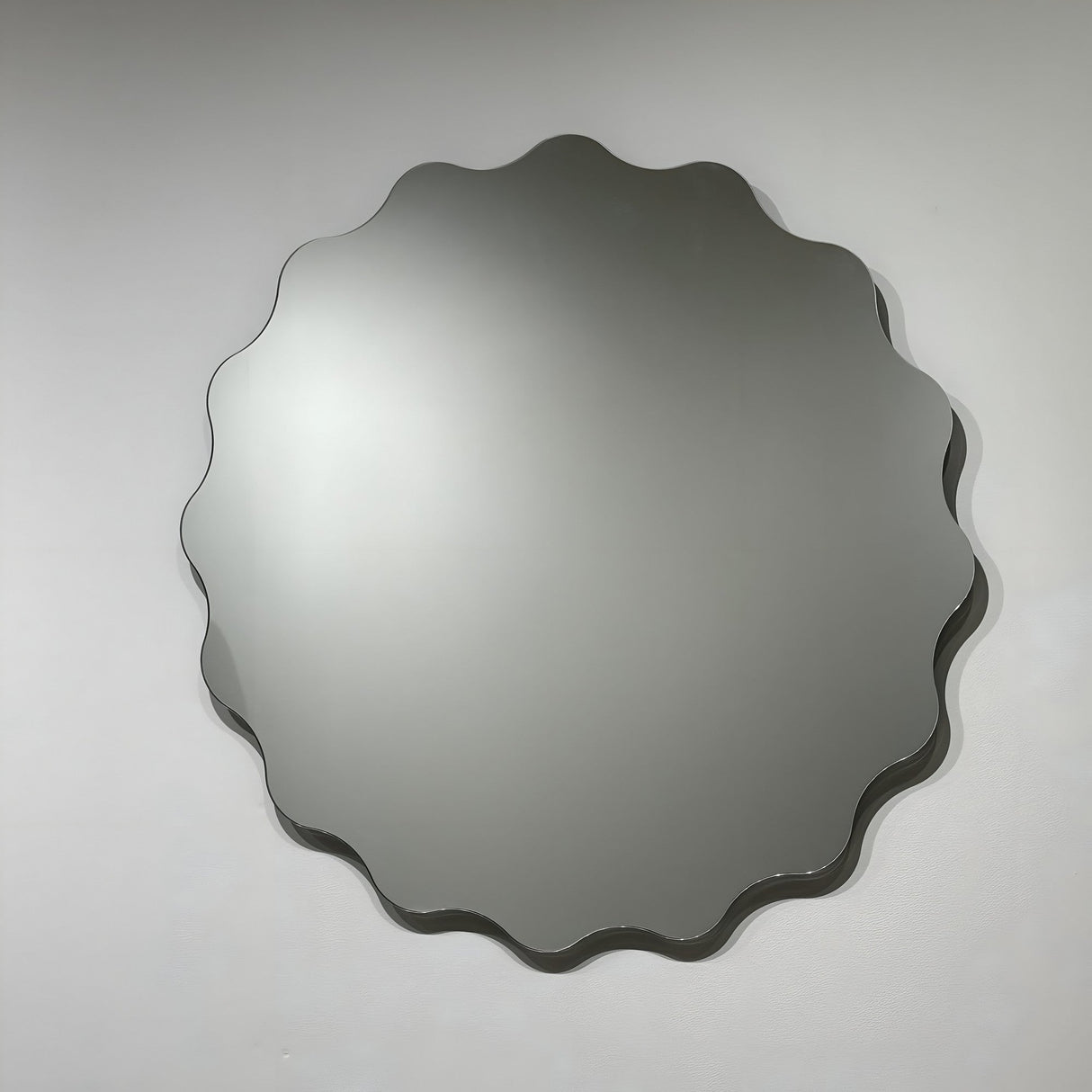 The Lumeo Circle Wall Mirror - Clear features a wavy, scalloped-edge frameless design that adds modern style to any space. Its smooth, reflective surface looks striking on a plain, light-colored wall.