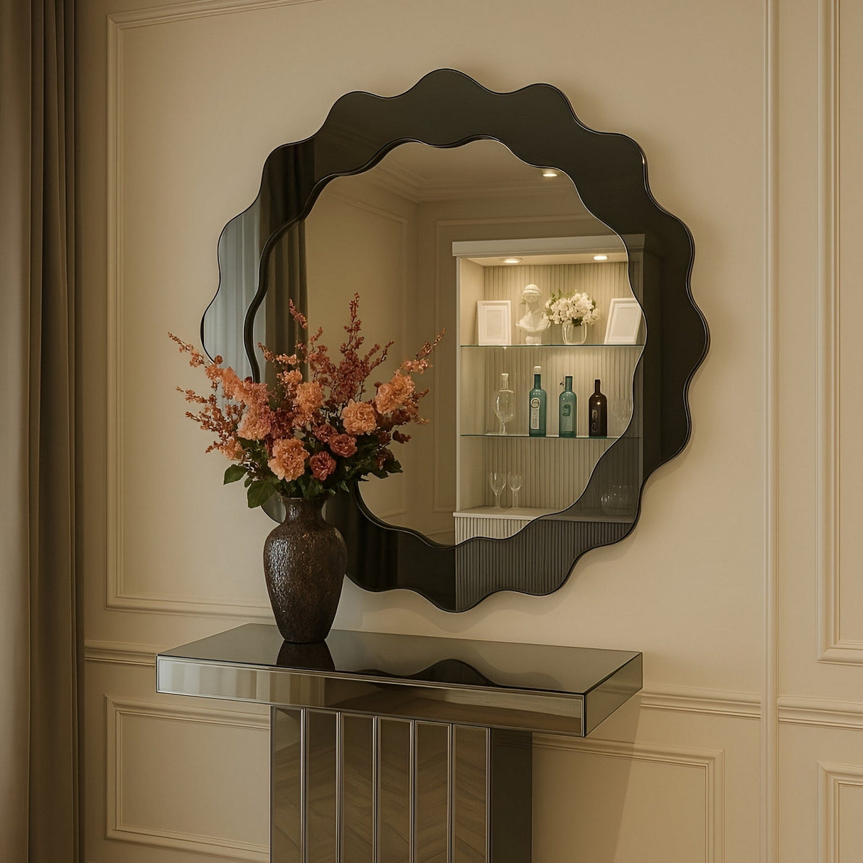 The Solstice Circle Wall Mirror - Grey, with its wavy edge design, hangs above a mirrored console table topped with a vase of pink and peach flowers, reflecting shelves lined with bottles and blooms behind.